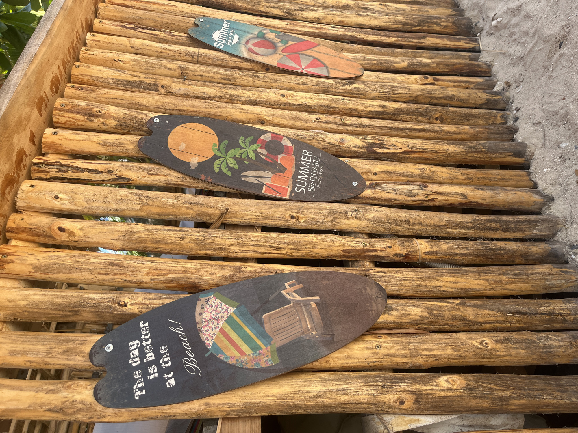 A ground-level shot of a walkway constructed of logs in Bac Natural Heritage Area Trang, Vietnam. Three decorative boards shaped like surfboards are affixed to the walkway. The top board is blue and features beach balls and the word "Summer Beach Party." The middle board is darker, displaying a sun, palm trees, a life preserver, beach chairs, and the words "Summer Beach Party Friday, 1 August." The bottom board features beach umbrellas, a lounge chair, and the phrase "The day is better at the Beach!" Screws or bolts are visible on the logs, indicating how the boards are attached. Sand is visible on the edge of the walkway, suggesting its proximity to a beach.