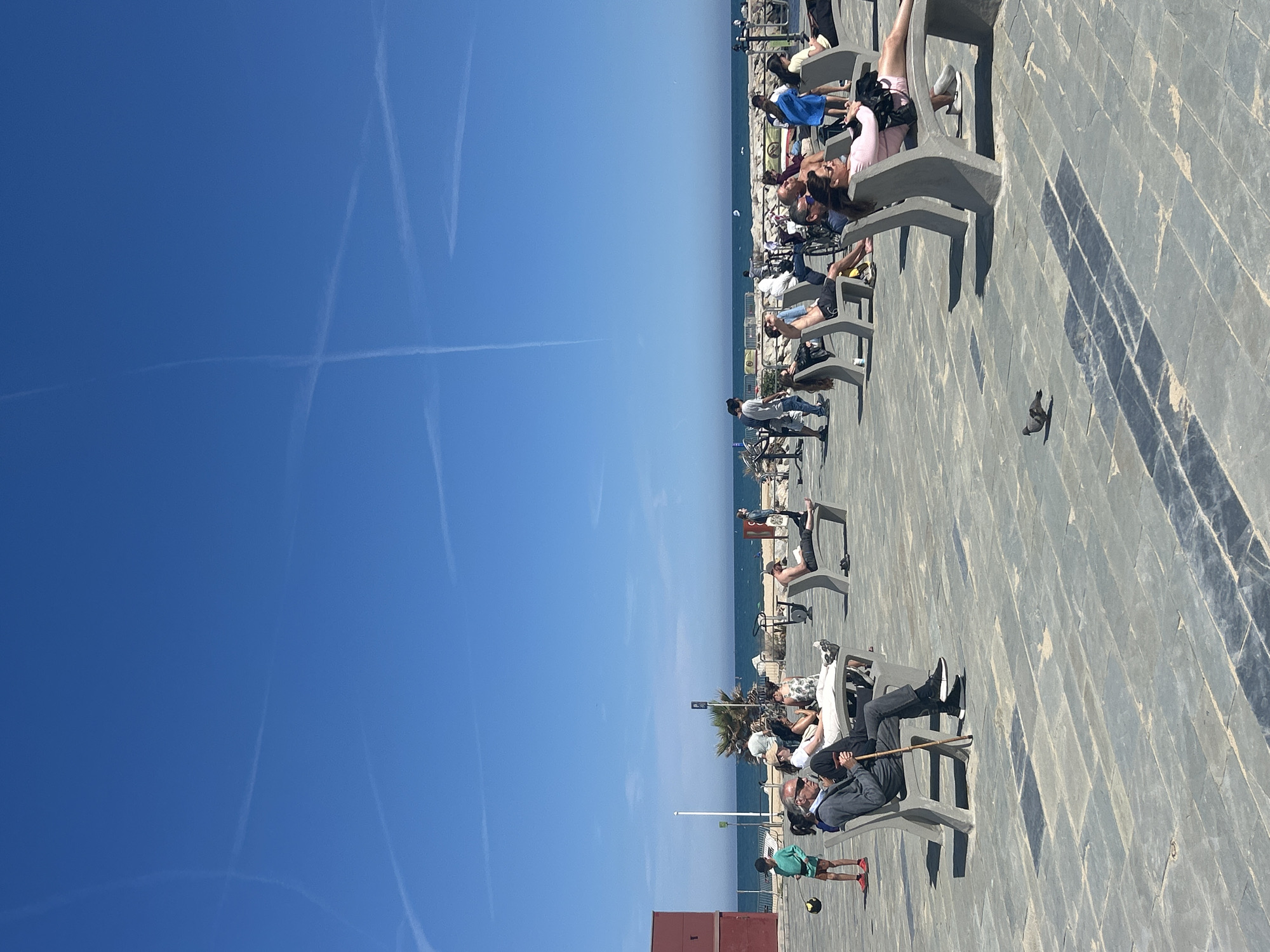 The image depicts a coastal scene in Barcelona, Spain. A series of modern concrete benches line a paved promenade, likely along a beach. Several people are sitting or lying on the benches, seemingly relaxing and enjoying the sunny weather. The sky is clear and blue, with prominent contrails from airplanes. In the background, the Mediterranean Sea is visible. A few palm trees and street lights punctuate the scene. A pigeon is depicted walking on the pavement.