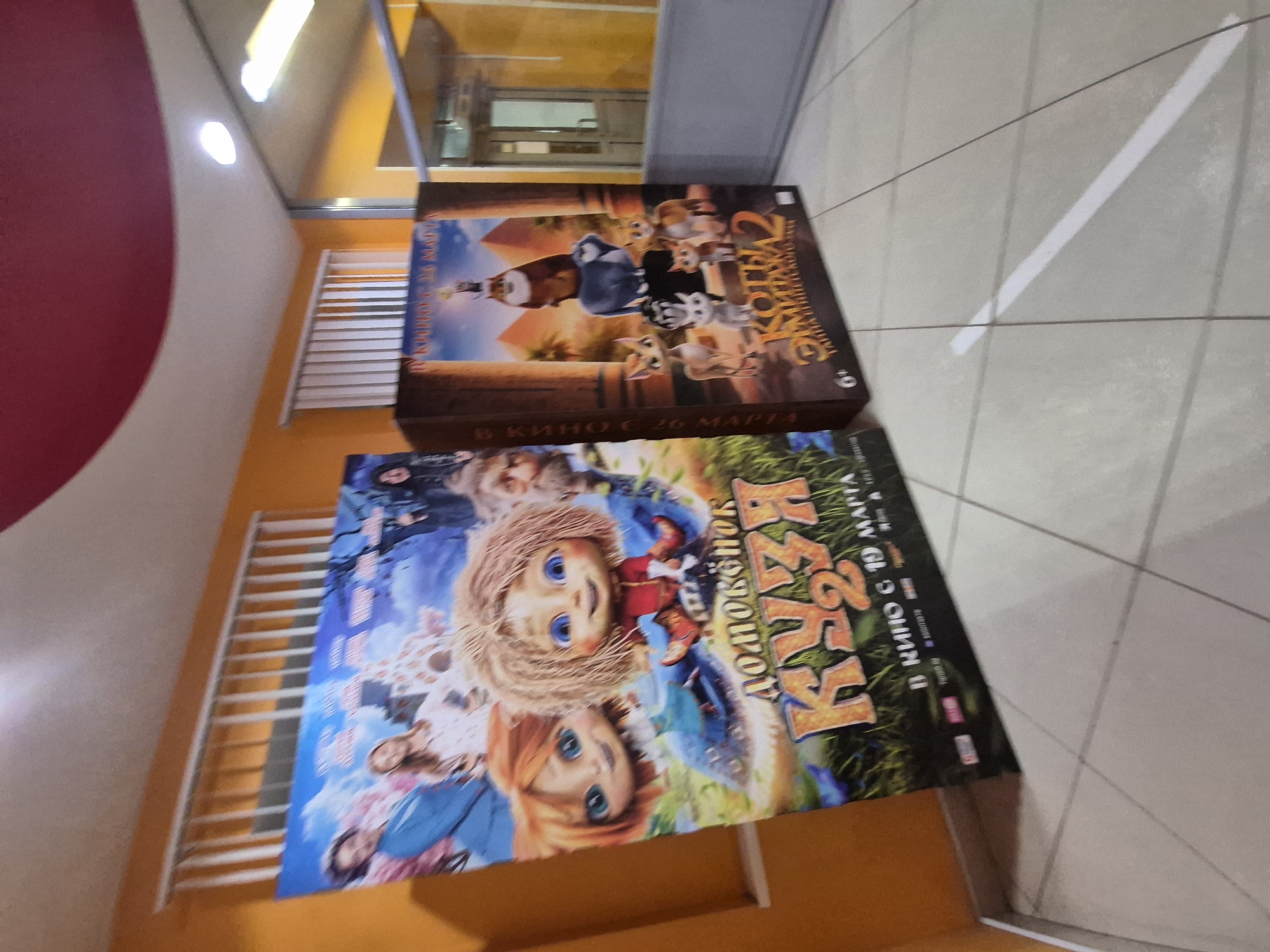 The image depicts two movie posters placed on a tiled floor in what appears to be an interior space, possibly a lobby or entrance area. The location is suggested to be in the city of Chaikovsky, Russia.
The top poster features the animated movie "Cats 2." The bottom poster features the animated movie "Домовой" (Domovoy) aka "Housewife 2." It indicates the release date "В кино с 19 марта" (In cinemas from March 19).
The tiled floor has a light gray tone with a diagonal white line marking. The walls are primarily orange and white with grilles running along them. The image is taken from an elevated perspective.