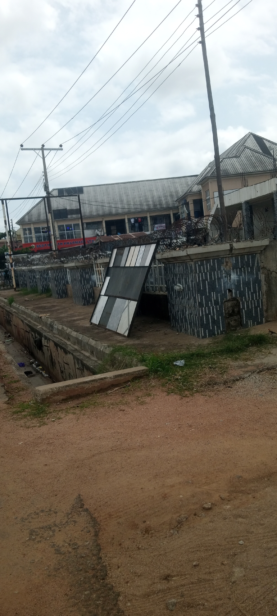 This image captures a street scene in Jalingo, Nigeria, likely in front of a commercial building. A prominent feature is a building with a corrugated metal roof and a light-colored facade, featuring multiple windows and what appears to be shops or offices. A sign reading "BEST HEALTH PHARMACY LTD" is visible on the building. In the foreground, a concrete drainage ditch runs along the side of the road, containing some visible debris. A low concrete barrier separates the ditch from the paved area. Adjacent to the drainage is a walled enclosure with a patterned facade and barbed wire on top, likely a security measure for the property. A large, multi-paneled object, possibly a large window or a sign, is propped up against this wall. A utility pole with multiple wires extends upwards into the cloudy sky. The ground is covered in reddish-brown earth and some sparse vegetation.