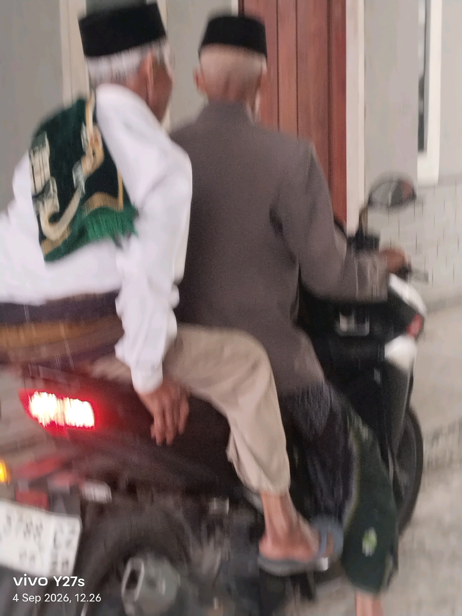 Two adult males, likely of Indonesian descent and wearing Islamic attire including peci caps, are seated on a dark-colored motorcycle. The individual in the foreground is wearing a white long-sleeved shirt and a green sash with gold trim over it. He is wearing light-colored pants and dark flip-flops. The second individual, positioned behind the first, is wearing a gray outfit and is driving the motorcycle. The motorcycle's red taillight is illuminated. The scene appears to be outdoors, possibly on a paved surface, with a building visible in the background. The lower left corner of the image shows a watermark that reads "vivo Y27s 4 Sep 2026, 12.26", suggesting the image was taken in Tlogo, Indonesia. The image is slightly blurred due to motion.