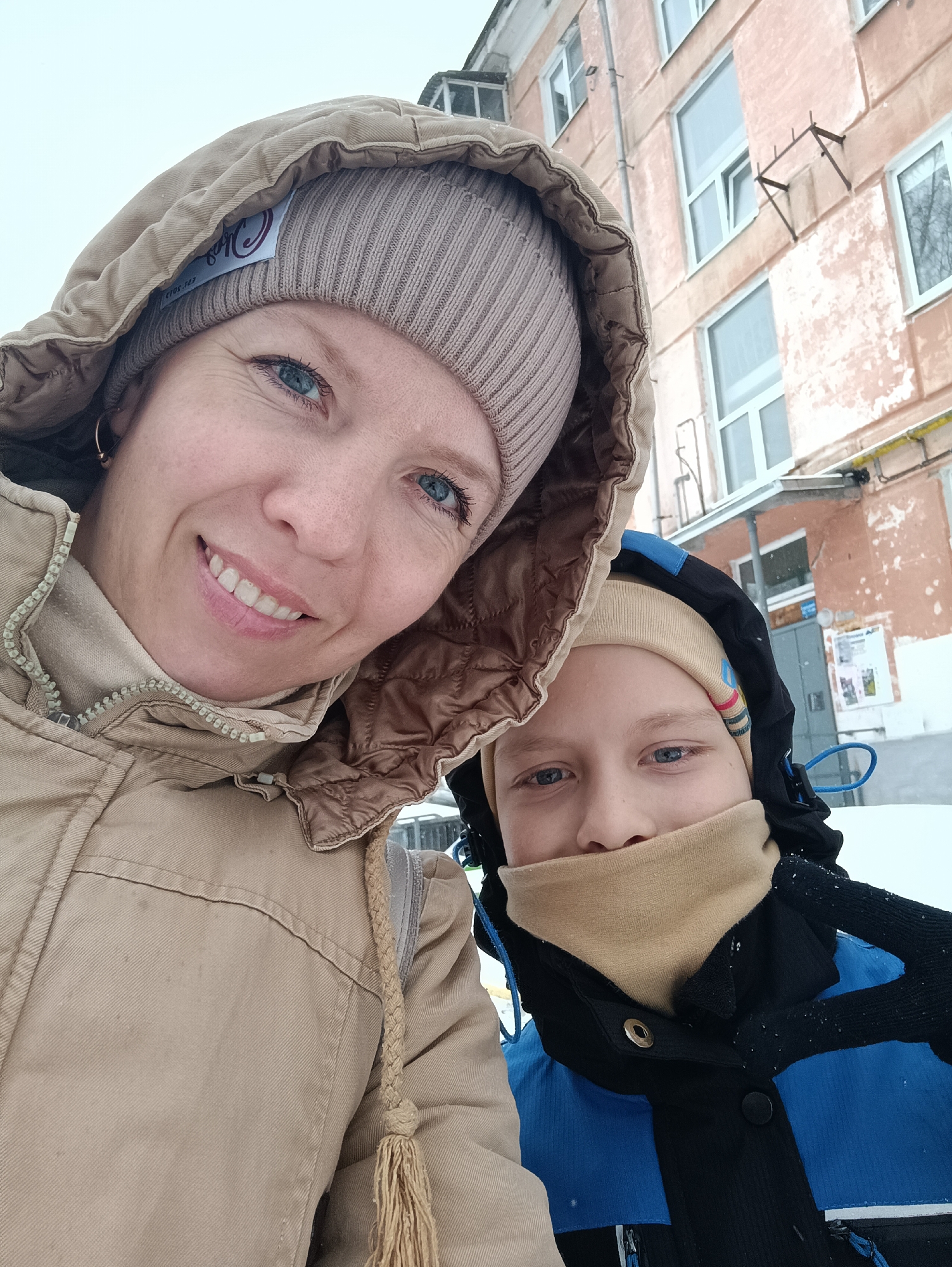 A woman and a child are pictured outdoors in the city of Chaikovsky, Russia. Both are wearing winter clothing. The woman, in the foreground, is fair-skinned with blue eyes and a warm smile. She wears a beige knit hat under the hood of her tan jacket, partially revealing its brown quilted lining. A beige braided cord hangs from her jacket. The child, standing slightly behind and to the side of the woman, has blue eyes and a tan-colored neck warmer that covers the lower half of their face. The child is wearing a black and blue winter jacket with a knit hat peeking out from under the hood. The background features a multi-story building with weathered peach-colored walls and white-framed windows. Snow is visible on the ground.