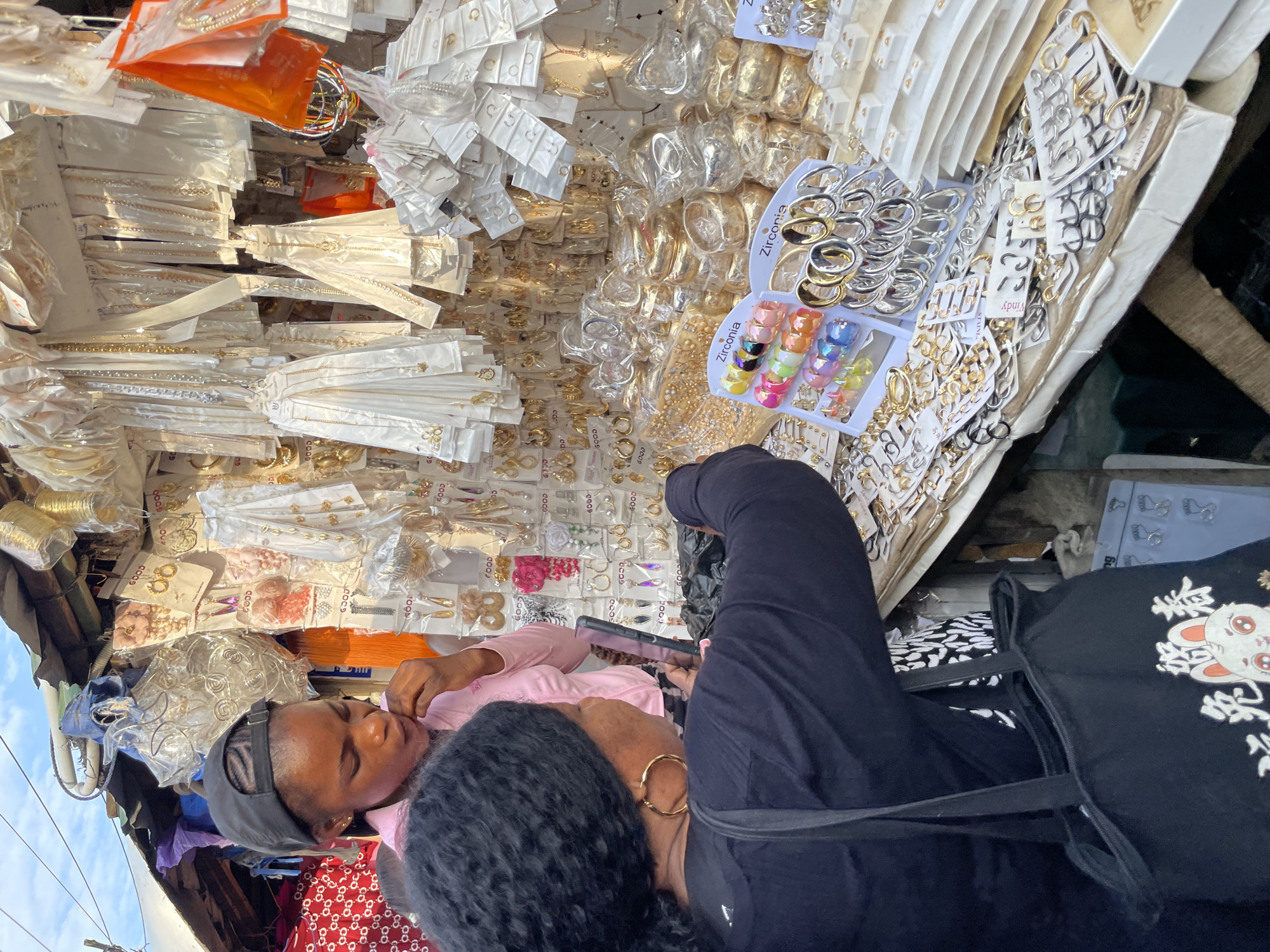 This media file shows a close-up view of a person looking at their phone in a market stall filled with jewelry. The stall is overflowing with various items, including necklaces, bracelets, earrings, and rings, many of which are packaged in clear plastic. The jewelry appears to be costume jewelry, with a mix of gold-toned and silver-toned pieces, as well as some colorful rings.
The person in the foreground is wearing a black top and black pants. They are holding a black smartphone in their right hand, and their attention is focused on the screen. Another person, wearing a pink shirt and a black cap, is partially visible in the lower left corner, also seemingly engaged with their phone.
The scene is outdoors, likely in a bustling market given the abundance of goods. The lighting suggests it could be daytime. The general atmosphere appears to be one of commerce and everyday life in a Nigerian setting, possibly Lagos, as indicated by the location context. Some text is visible on the packaging, including "GOOD" and "Zirconia," indicating brands or material types. The overall impression is one of a busy marketplace where people are browsing or working.