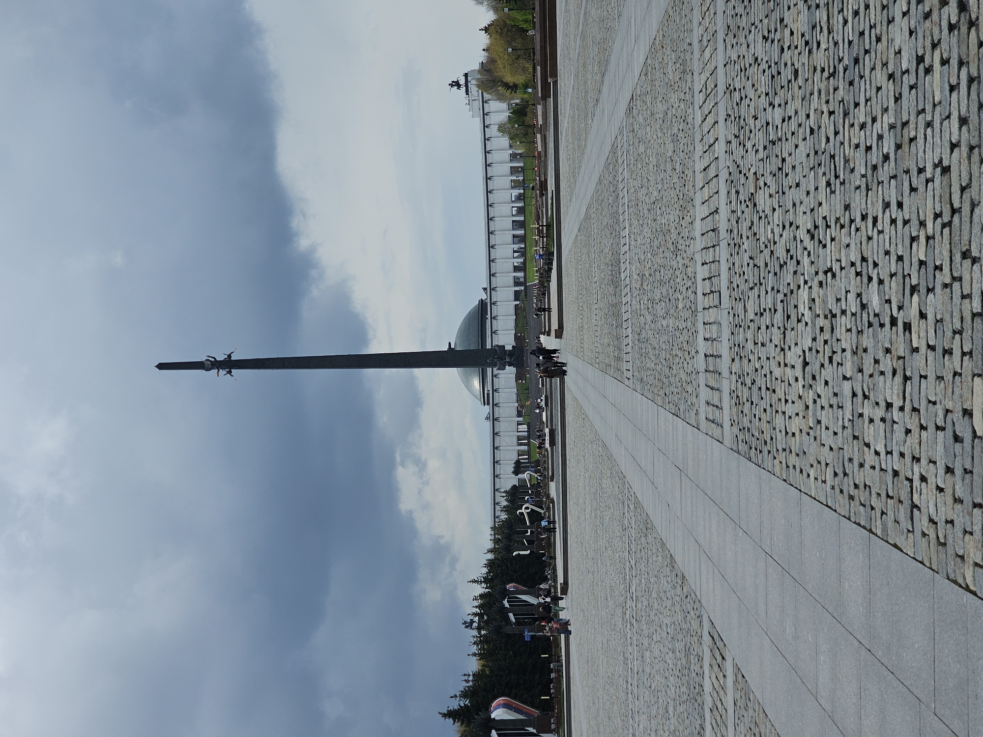 The image shows the Victory Monument in Victory Park, Moscow. The monument is a tall obelisk with a statue of Saint George slaying the dragon on top. The monument is surrounded by a large plaza paved with stone. In the background is the Central Museum of the Great Patriotic War. Rows of memorials are visible along the sides of the plaza. The sky is overcast. There are a few people in the background.