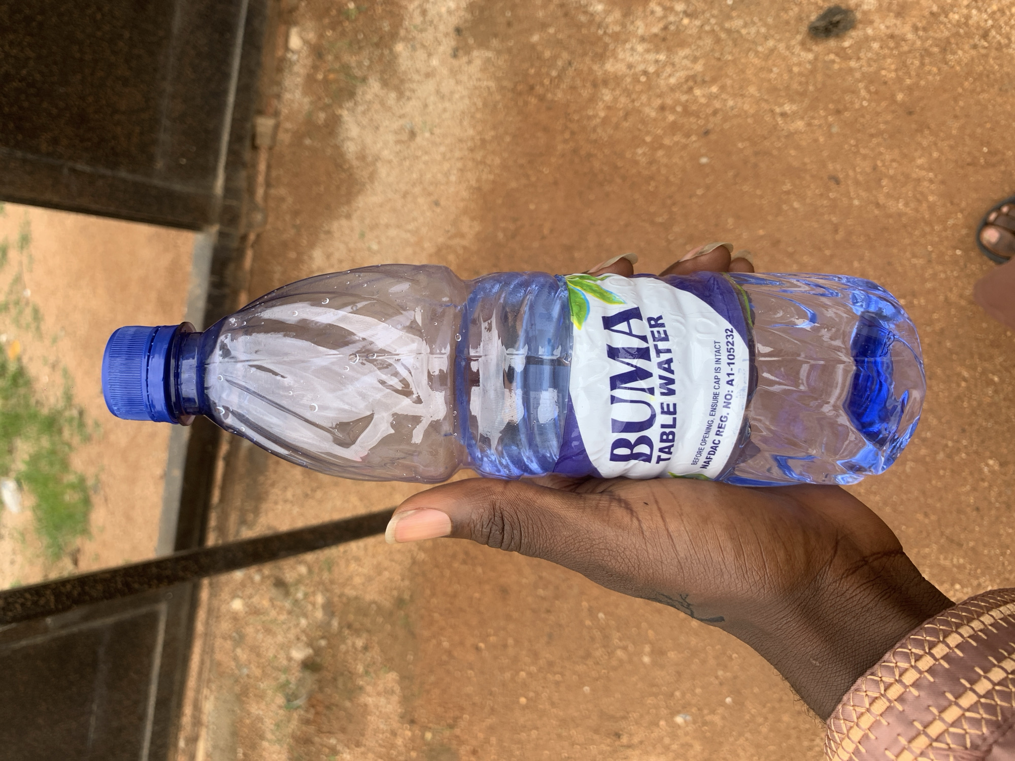 A person's hand holds a clear plastic bottle of BUMA Table Water. The bottle has a blue cap and a white label with purple text that reads "BUMA TABLE WATER". Below this, smaller text states "BEFORE OPENING, ENSURE CAP IS INTACT" and "NAFDAC REG. NO: A1-105232". The bottle contains clear liquid with a blue tint. The background appears to be an outdoor, unpaved area with reddish-brown soil and scattered green vegetation. A dark metal structure, possibly a fence or gate, is visible in the upper left of the image. The scene is likely located in Howayi, Nigeria.