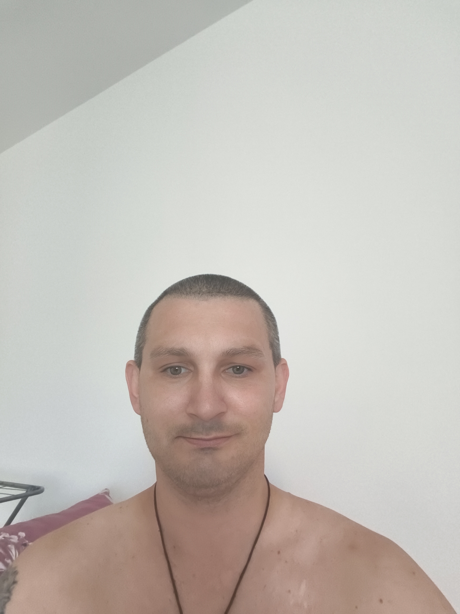 A man with a buzz cut and light stubble is shown from the shoulders up, looking directly at the camera. He has light skin and light-colored eyes. He is wearing a thin, dark cord necklace. The background is a plain, lightly textured white wall, with a diagonal shadow suggesting a change in light from a source not visible in the frame. The man appears to be indoors, possibly in a residential setting. No specific location in Wolgast, Germany can be determined from the image.