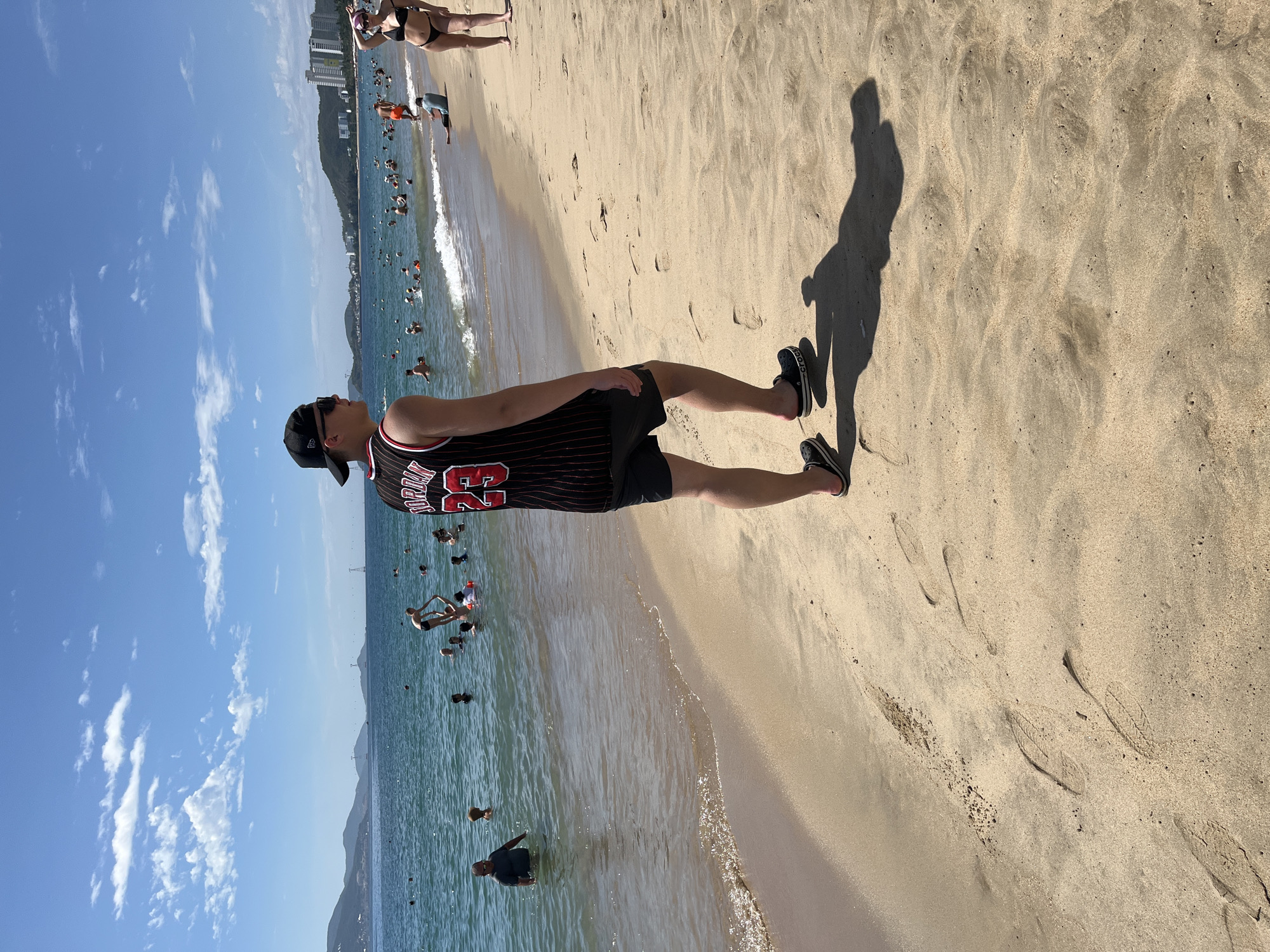 In Nha Trang, Vietnam, on a sunny day, people are enjoying themselves on the beach and in the water. The sand is light brown, with footprints visible in some areas. The ocean is a clear blue, with small waves lapping at the shore. In the background, a city skyline with buildings can be seen along the coast, and mountains rise in the distance.
In the foreground, a young man is walking on the beach, seen from the back and right side. He is wearing a black baseball cap, sunglasses, a black and red striped basketball jersey with the number 23, and black shorts. He is also wearing black Crocs-style sandals. His shadow is cast on the sand behind him.
Further down the beach, a woman in a black bikini is posing with her hand on her head. Other beachgoers are scattered across the sand and in the water, swimming, playing, or relaxing. The water is populated with many people, and there is a sense of activity and leisure. The sky is a vibrant blue, with scattered white clouds. The overall scene depicts a lively beach atmosphere in Nha Trang.