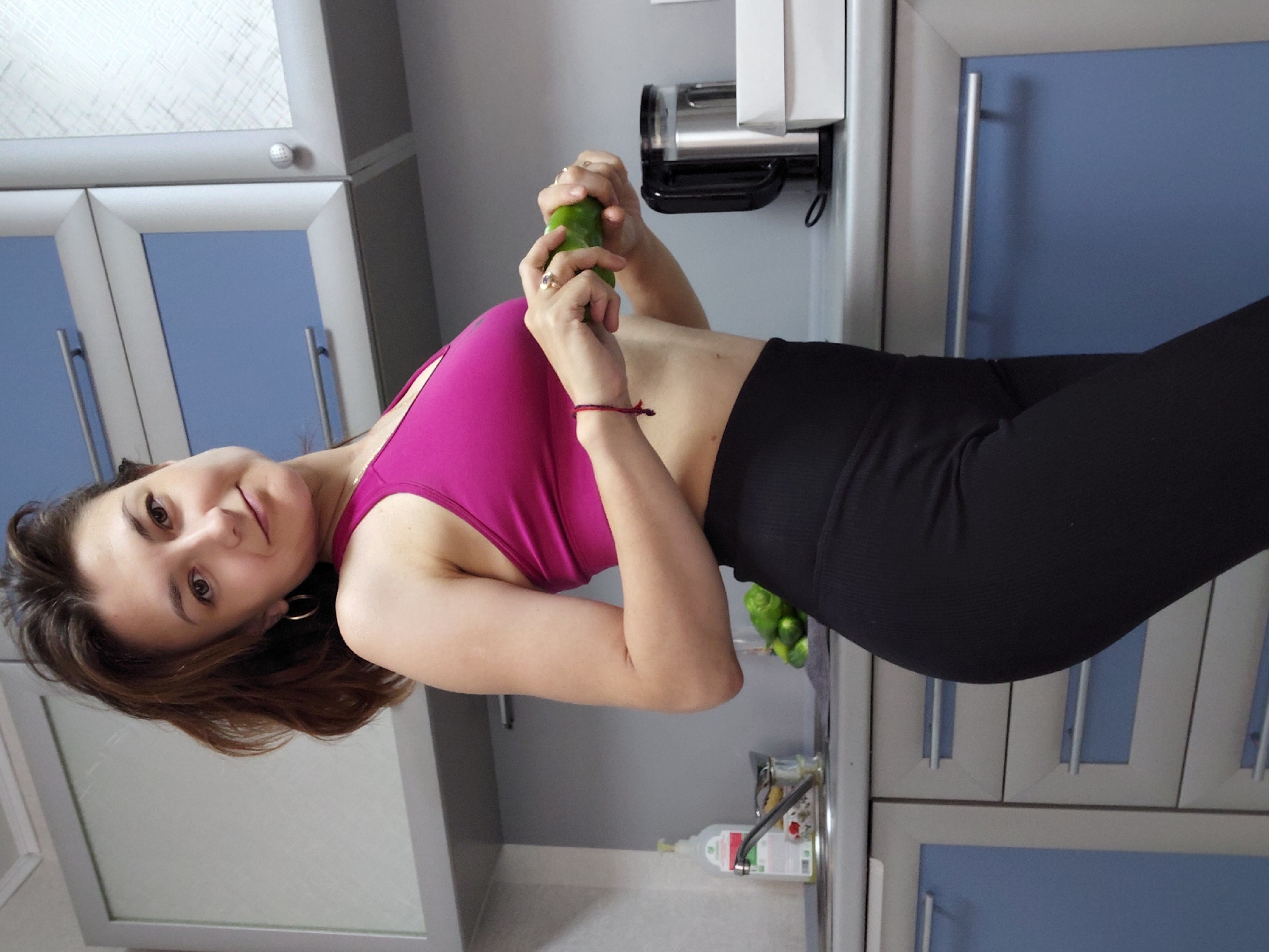 A woman stands in a kitchen. She is wearing a pink sports bra and black leggings. She is holding a cucumber in her hands. The kitchen has gray cabinets and blue doors. There is a sink, a faucet, and a bottle of dish soap on the counter. There is also a kettle and a white box on the counter. In the background, there are some cucumbers in a bowl. The woman has brown hair and is wearing hoop earrings. She is smiling at the camera. This image was taken in Magnitogorsk, Russia.