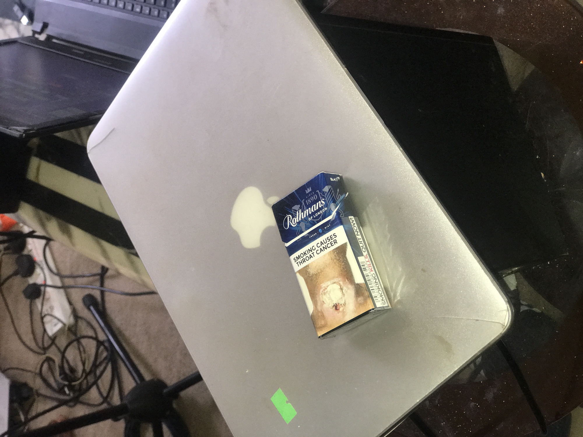A pack of Rothmans cigarettes lies on top of a silver MacBook laptop. The pack features a graphic depicting throat cancer and warnings about smoking. The laptop is positioned on a table with a dark surface and various electronic accessories and wires are scattered nearby on a carpeted floor.