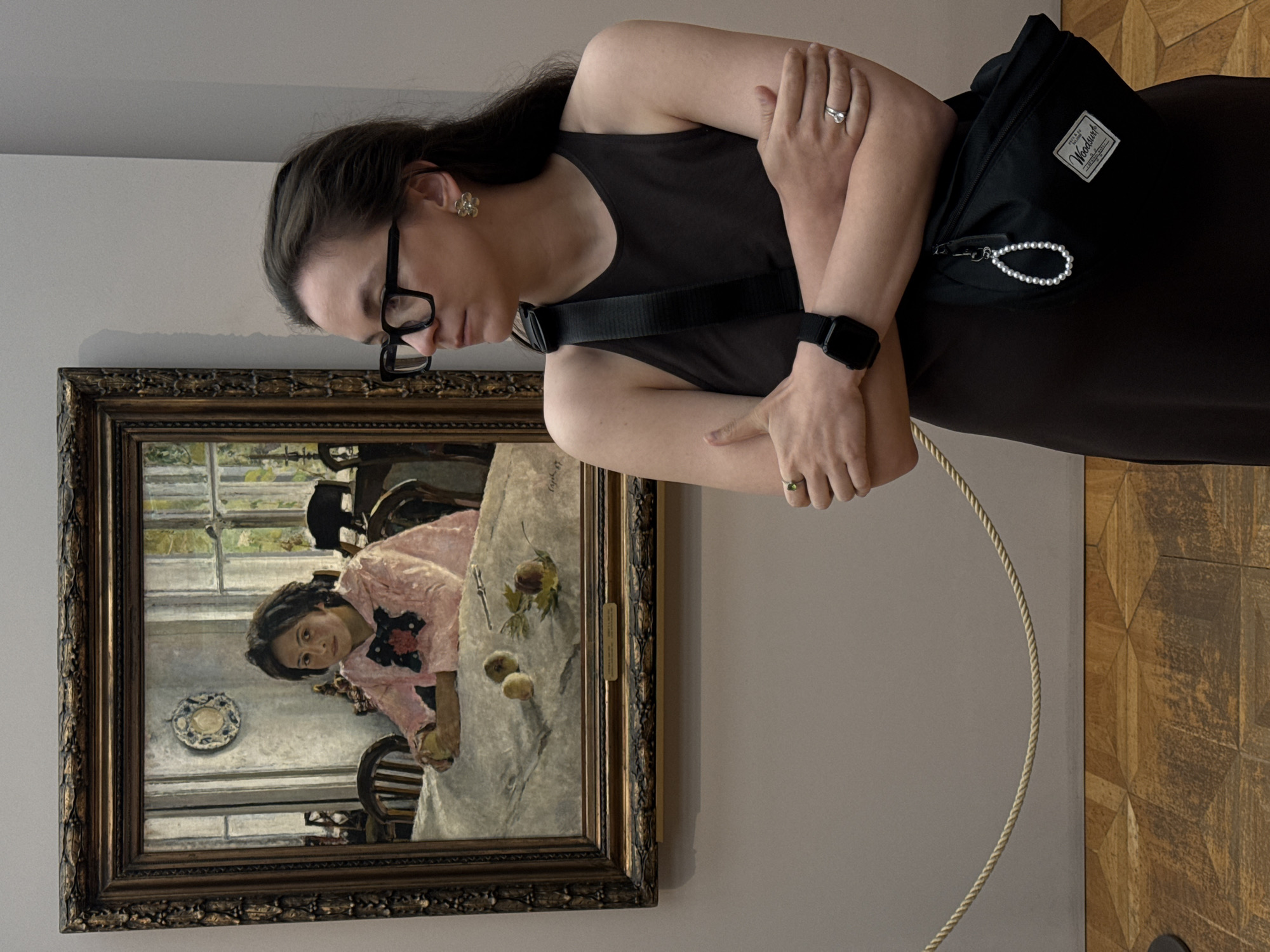 A woman wearing glasses and a black dress with a Herschel bag is standing in front of a painting on a wall. She has a smart watch on her left wrist, and rings on both hands. The painting is framed and depicts a woman sitting at a table with fruit. The background of the painting shows windows and chairs. The wall is light gray. The floor is wood with a geometric pattern. The woman is looking at the painting. A rope is extending up from the bottom. The location is the Tretyakov Gallery in Moscow, Russia.