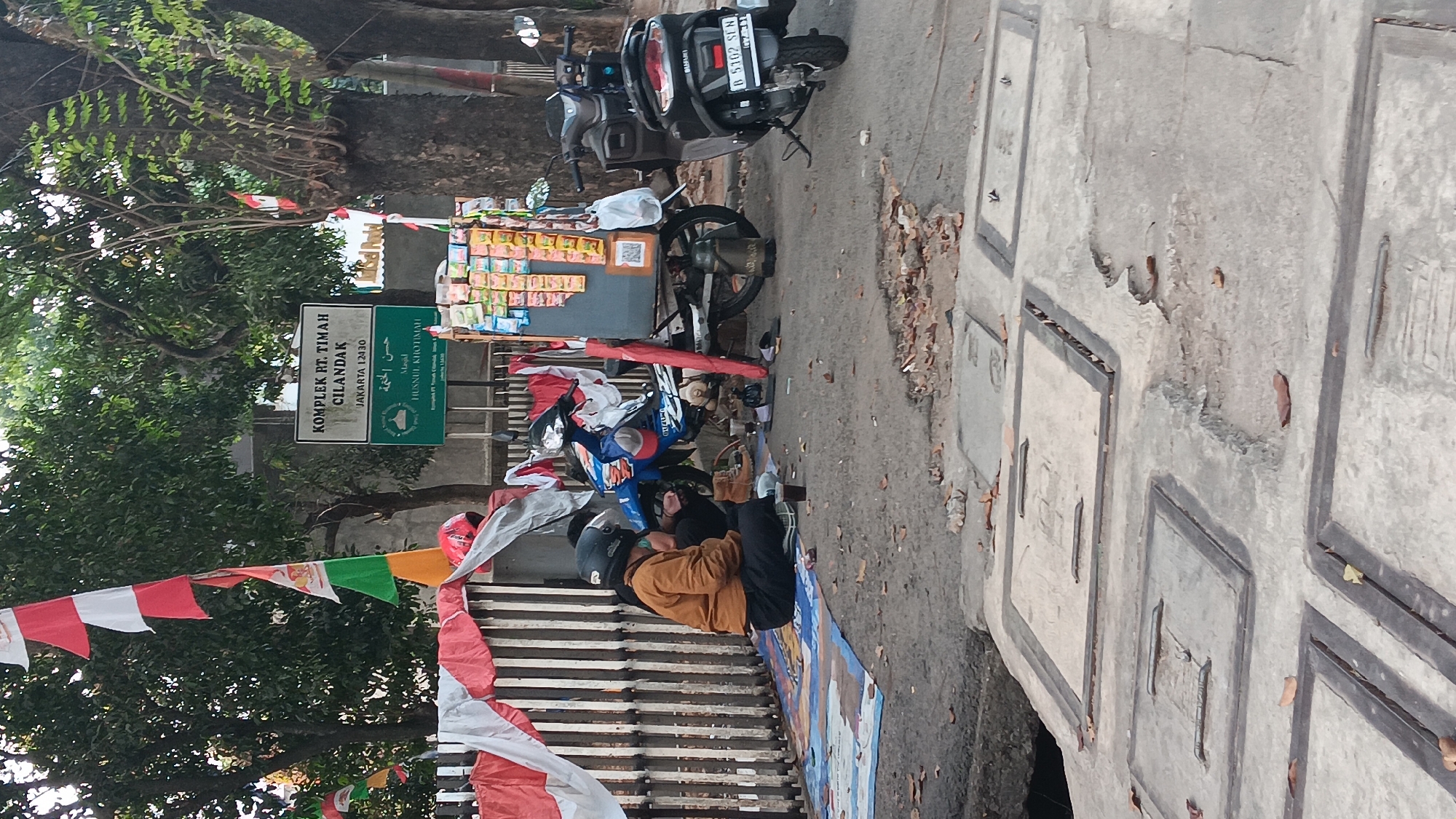 The image depicts a street scene in South Jakarta, Indonesia, likely near Kompleks RT. Timah, Cilandak, Jakarta 12430, as indicated by a sign. In the foreground, a person wearing an orange jacket and a black helmet sits on a blue mat on the ground, next to a small table with drinks and a gas canister. Two motorcycles are parked nearby. One is a blue and white Suzuki, and the other is a dark scooter with a license plate reading "B 5102 SEN". Behind them, a street vendor's cart is laden with packaged goods. The street is paved, and several manhole covers are visible in the foreground. Red and white Indonesian flags are draped over a fence in the background, suggesting a national celebration or event. The overall environment appears to be an urban street setting with trees and foliage.