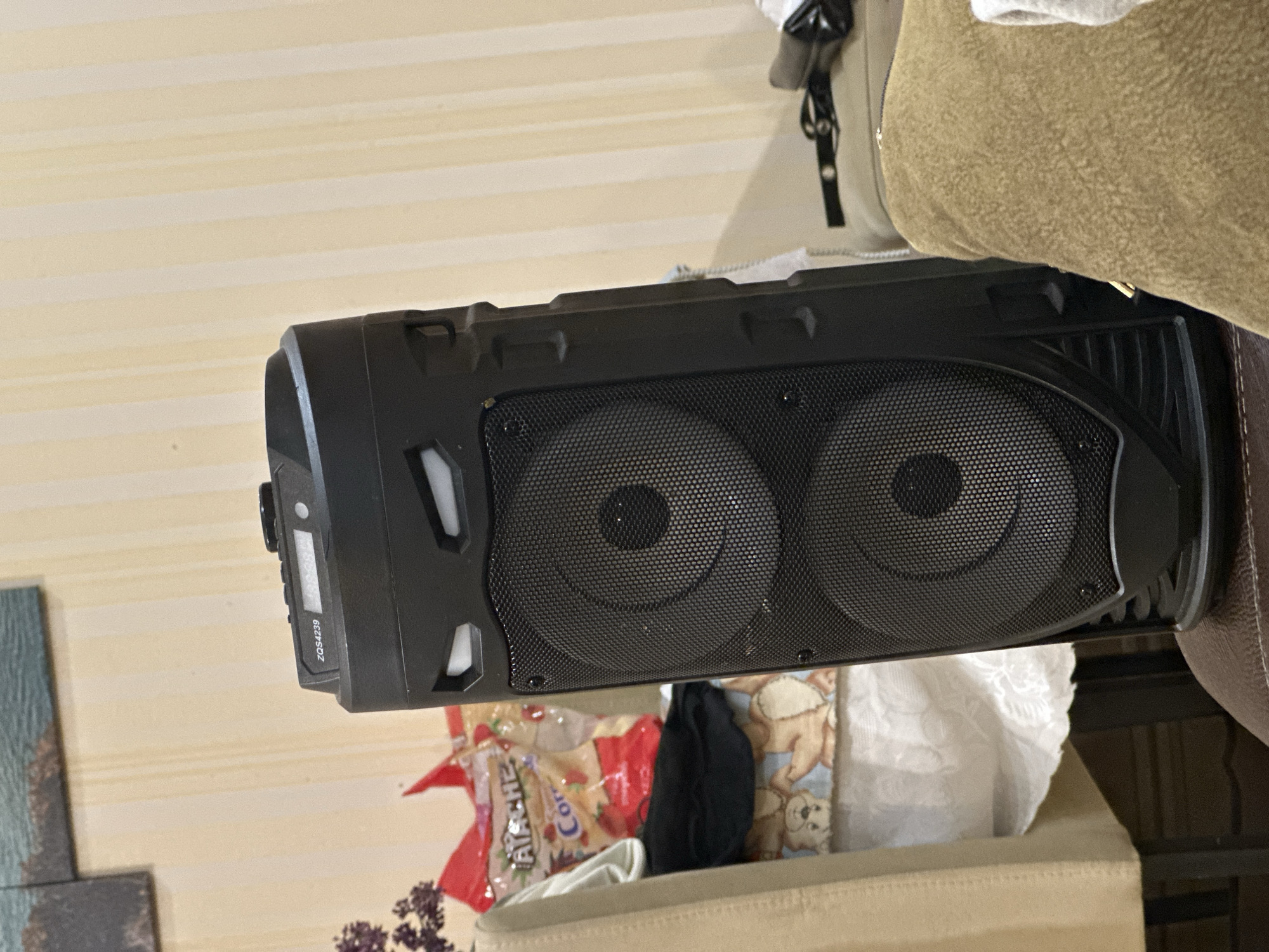 The image shows a black portable speaker positioned vertically, resting on or next to a piece of upholstered furniture, possibly a couch or chair. The speaker features two prominent speaker grilles on its front and controls on its top/side. Visible text on the speaker reads "ZQS4239".
The background consists of a cream-colored wall with distinct horizontal stripes. To the left, a portion of a framed artwork or canvas is visible. Below the speaker, several items are placed on a lighter-colored surface, including a red and white bag with the text "ATASHE" and "Cor" partially legible, a piece of black fabric, and a light blue patterned fabric showing a teddy bear design. A white, lace-like fabric is also visible on the right.
The setting appears to be an indoor room, likely a living area. There is no specific activity taking place; the speaker is stationary. The lighting suggests ambient indoor light. The image itself is rotated 90 degrees clockwise.