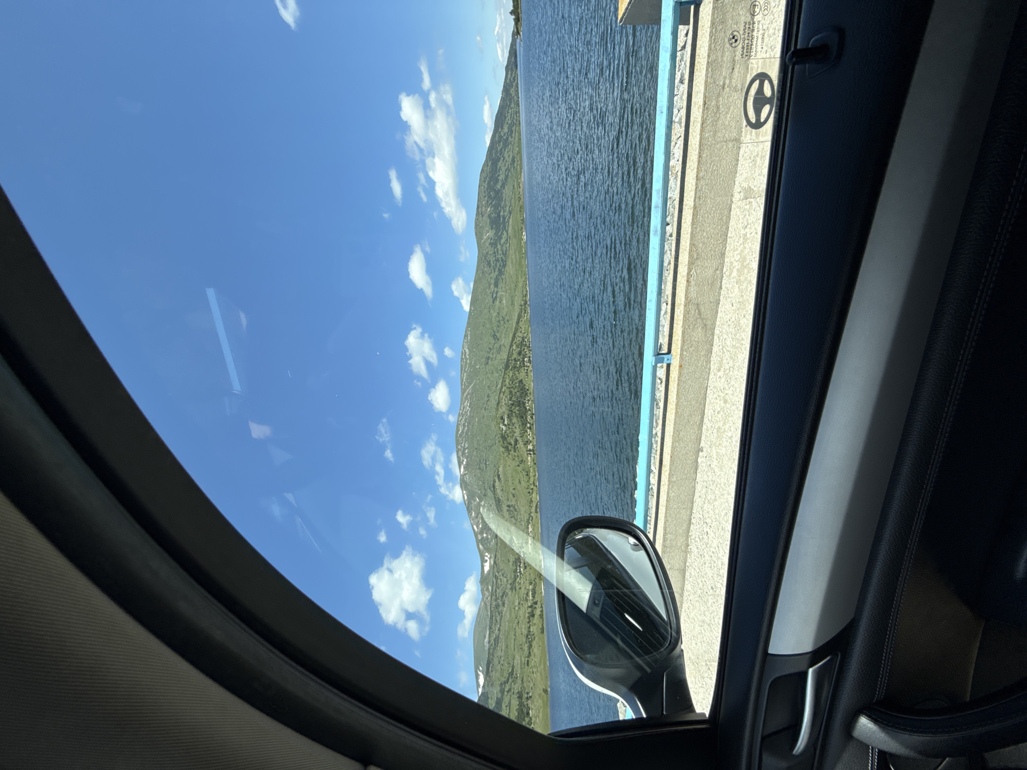 The image captures a scenic view from inside a car, looking out the passenger window towards a large body of water and a mountain range under a clear blue sky with scattered clouds. The water appears to be a lake or reservoir, with its surface rippling slightly. The mountains are green and rolling, with patches of snow visible on their peaks. A road or pathway with a light blue railing runs along the edge of the water. The interior of the car is visible in the foreground, including the car door, a side mirror, and the car's roof lining. The overall impression is a picturesque natural landscape seen during a car journey. The specific location in the city of Smolevo, Bulgaria, is not evident from the visual elements within the image.