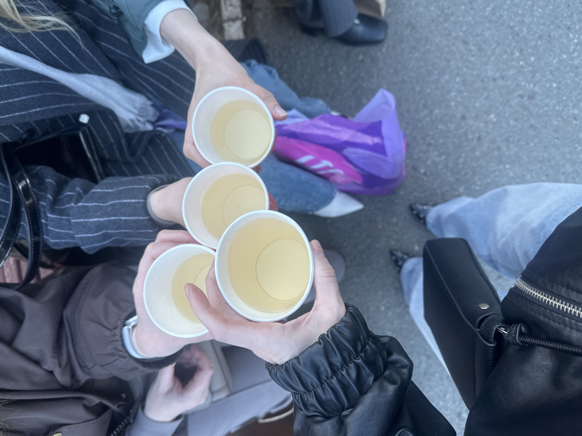Four people are holding paper cups filled with a light-colored liquid, likely tea or juice, in what appears to be a celebratory toast. They are standing outdoors on a paved surface, presumably a street or sidewalk. The individuals are wearing various jackets and coats, suggesting a cool climate. The scene may be located in a city, potentially Izhevsk, Russia, given the request context, but without additional visual information, this can't be verified.