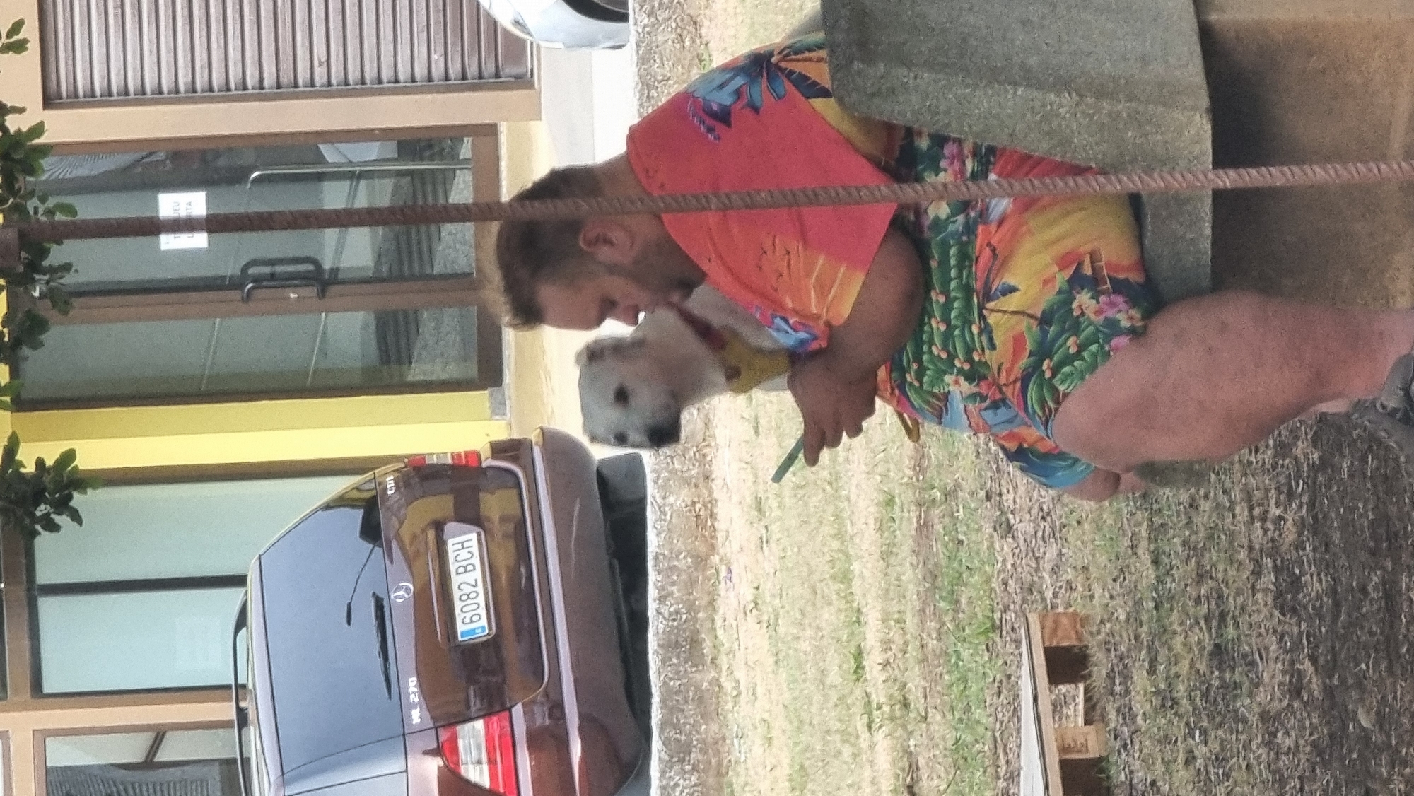 A man is sitting on a stone bench with a white dog sitting beside him, both looking at a smartphone. The man is wearing a vibrant, tropical-patterned shirt and shorts. The dog is wearing a yellow harness. Behind them, a dark red Mercedes-Benz SUV is parked. The scene appears to be outdoors, with a grassy area and a building with large windows and a yellow facade in the background. The setting is likely a public space in Sant Joan Les Fonts, Spain, given the context provided, possibly a park or a plaza.