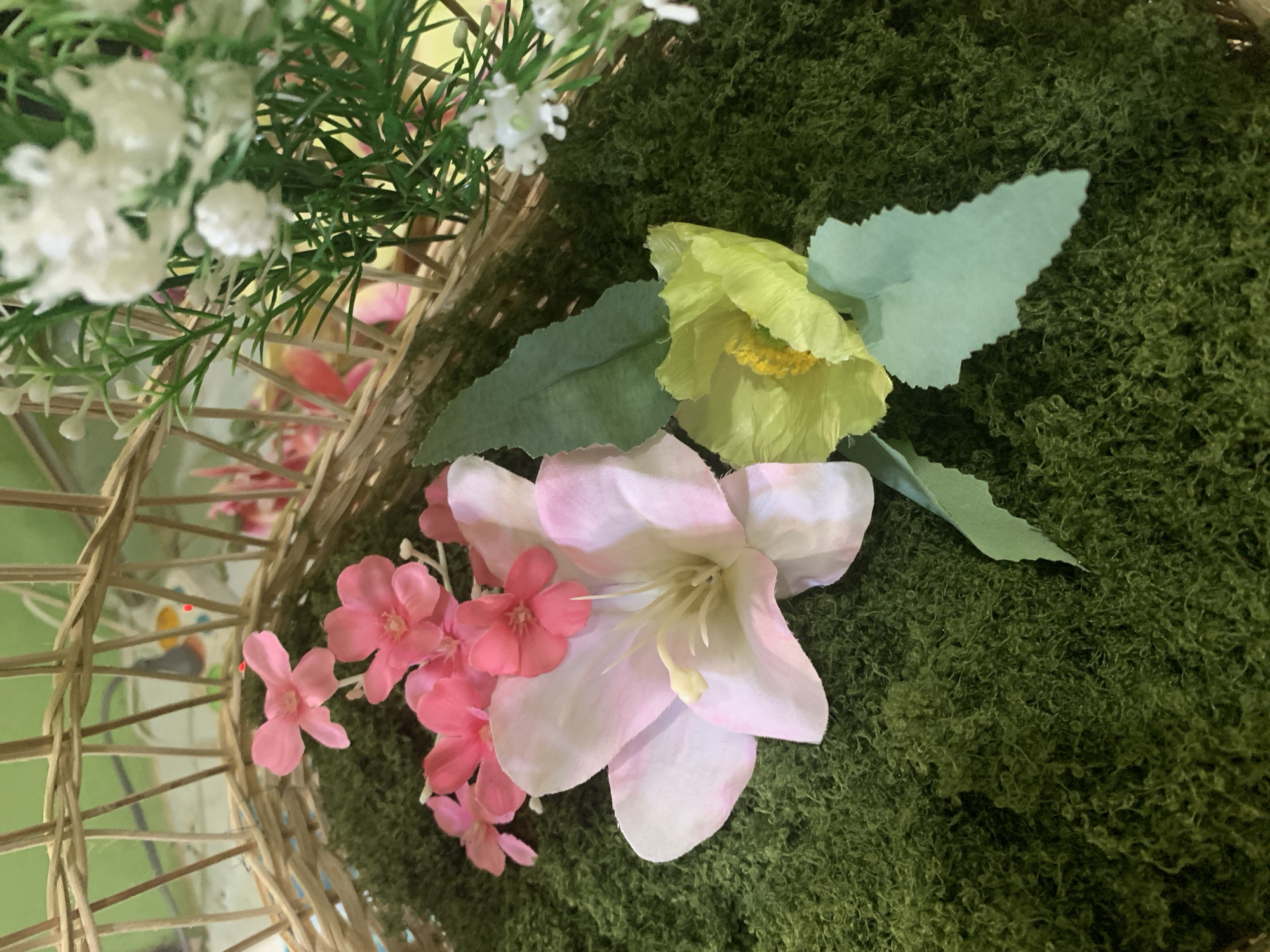 This is a close-up, top-down shot of a wicker basket filled with artificial flora. The base of the basket is covered in a thick layer of green, moss-like material, providing a lush, earthy texture. Scattered across this green base are several artificial flowers.
In the foreground, a large, pale pink lily-like flower is prominent, its delicate petals tinged with white and a soft yellow stamen at its center. To its right, a smaller, bright yellow poppy-like flower with ruffled petals and a dark yellow center is visible.
Behind these, a cluster of small, vibrant pink flowers are arranged, some overlapping each other. Further back, a hint of greenery from other artificial plants can be seen, along with parts of the wicker basket's structure, which appears to be woven from light-colored reeds. The background is softly blurred, suggesting a shallow depth of field, with hints of green and yellow, possibly from more foliage or a decorative backdrop.
The image is a still life, focusing on the arrangement of artificial flowers and greenery within a decorative basket. There are no people, actions, or interactions depicted. The scene is static and purely visual.
Without any external context or landmarks, it is impossible to determine the specific location within Kuala Pembuang, Indonesia. This image depicts a decorative arrangement that could be found in various indoor settings, such as a home, shop, or event venue.