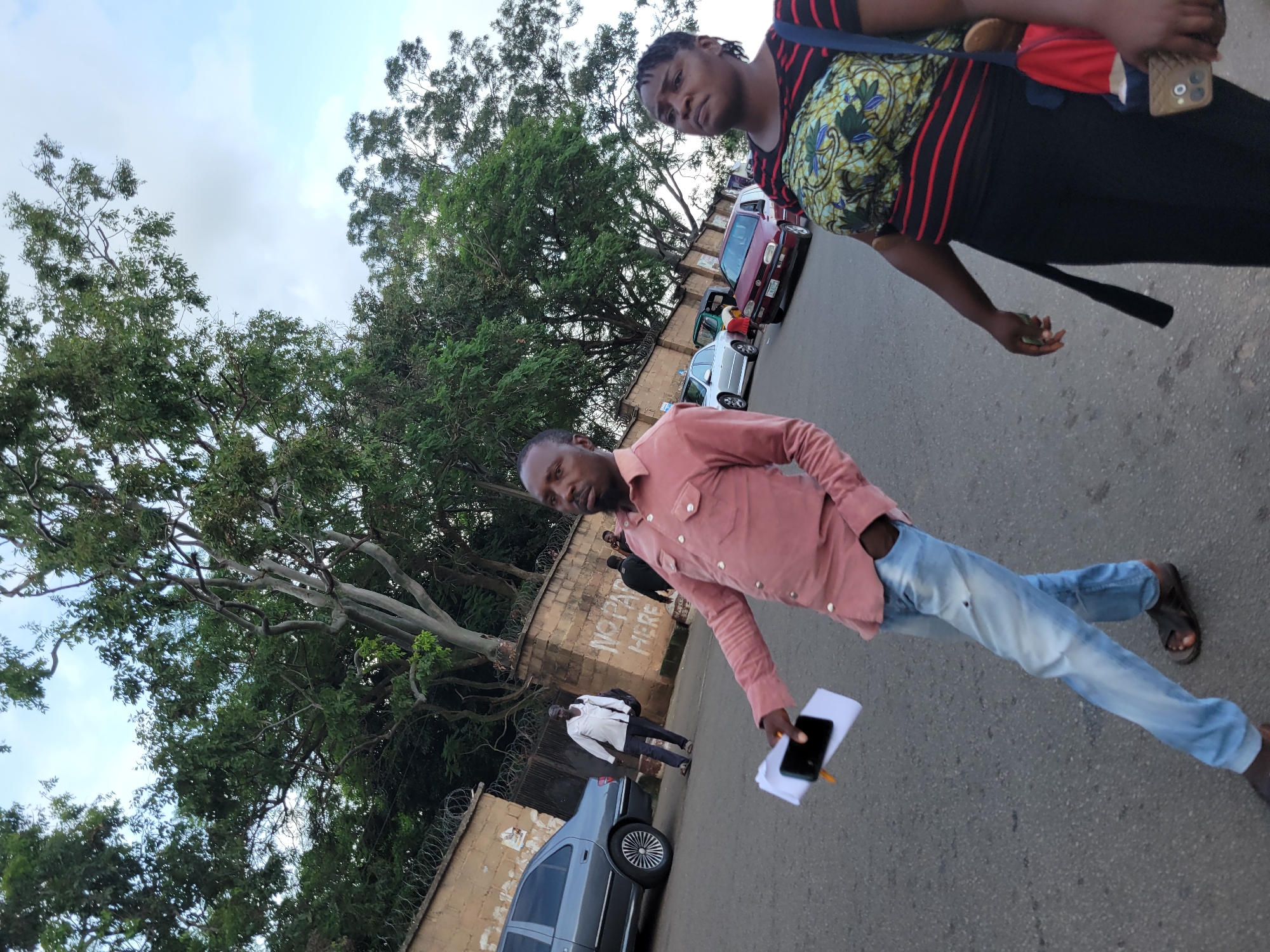 In the city of Jos, Nigeria, on a paved street, a man in a pink collared shirt and light blue jeans walks forward, holding papers and a phone. He wears dark sandals. To his right, a woman in a black and red striped shirt with a patterned fabric around her chest and black pants walks alongside him. She carries a red bag and a phone. In the background, a wall topped with barbed wire borders the street, with trees and foliage behind it. Several cars are parked on the street, including a grey sedan, a red hatchback, and a small green vehicle. Another man in a white shirt and dark pants walks further down the street, carrying a bag over his shoulder. The sky is partly cloudy.
