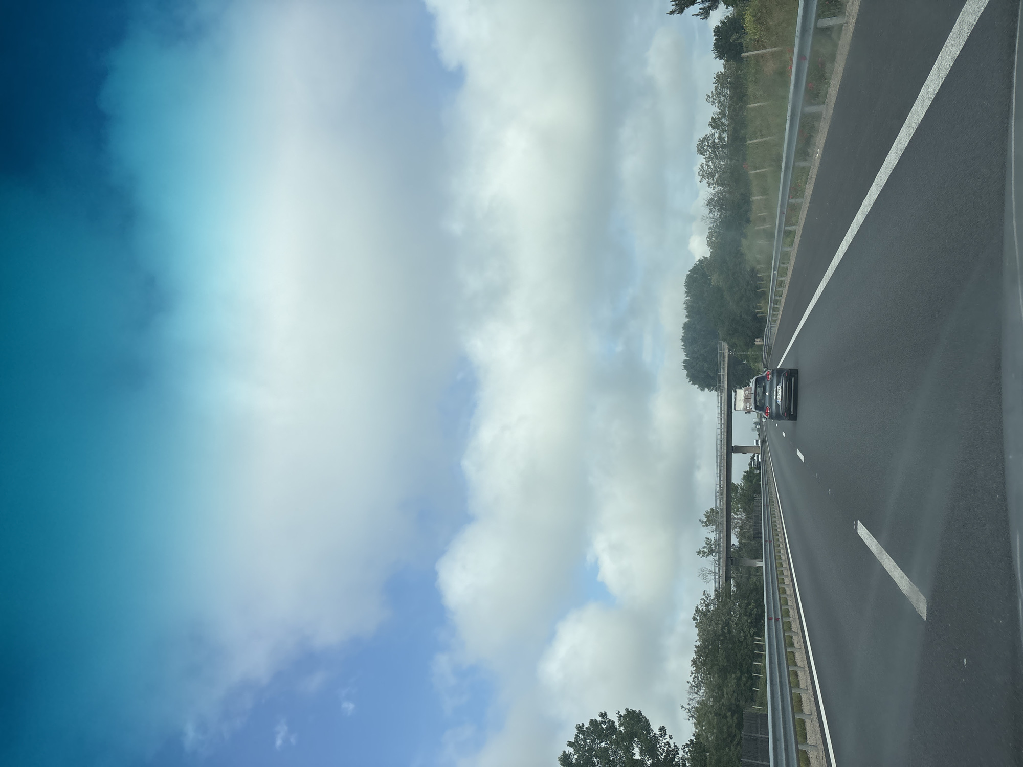 This media file captures a view from a moving vehicle on a multi-lane highway during the daytime, specifically in the Gorni Bogrov area of Bulgaria. The image is oriented vertically but depicts a horizontally stretching road.
The scene shows a modern asphalt highway with clear white lane markings. The vehicle from which the picture is taken appears to be in the rightmost lane. Further ahead, a dark-colored car, possibly a hatchback or small SUV, is visible in the same lane. The highway is bordered by a metal guardrail on the left, behind which are dense green trees and other foliage, suggesting a semi-rural or natural environment alongside the road. In the distance, an overhead structure, likely an overpass or bridge made of concrete or metal, spans across the highway.
The sky occupies the upper portion of the image. It is partly cloudy, with scattered white cumulus clouds against a varying blue background. There's a noticeable bright area in the upper left of the sky, indicating direct sunlight or a strong glare. The overall weather appears clear and bright.
No discernible people are visible, and no legible text can be identified on signs, vehicles, or other elements in the image. The activity taking place is typical highway travel.