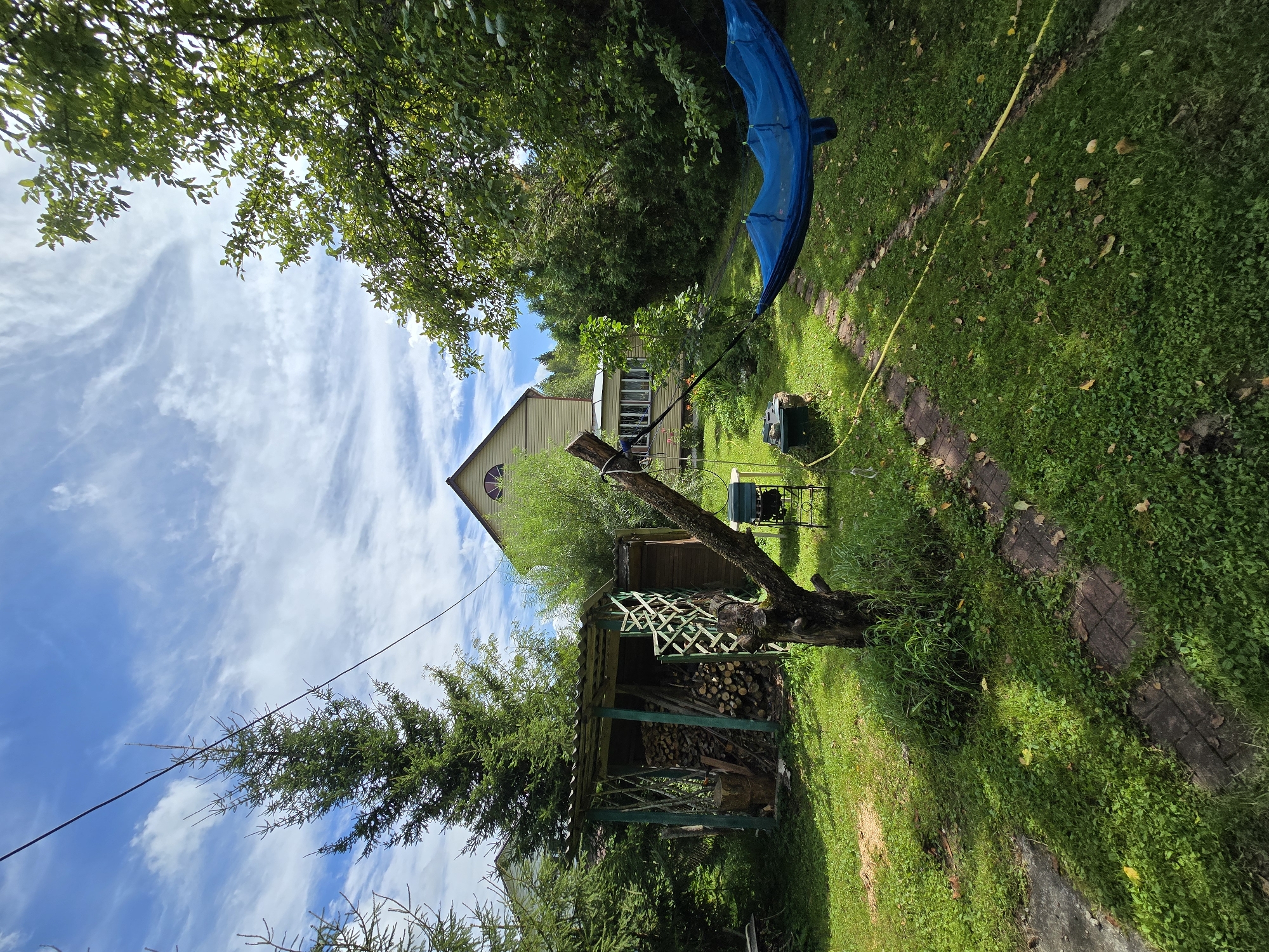 The image depicts a backyard scene in Lobkovo, Russia, on a sunny day with scattered clouds. A two-story house with a light yellow exterior and a semicircular window is visible in the background, surrounded by lush green trees. In the foreground, a blue hammock is suspended between two points, one appearing to be a tree trunk. A path made of brick pavers winds through the grassy yard. To the left, a wooden structure, possibly a woodshed, is filled with logs. Various garden tools and equipment are scattered throughout the yard. The overall impression is one of a tranquil, rural setting.