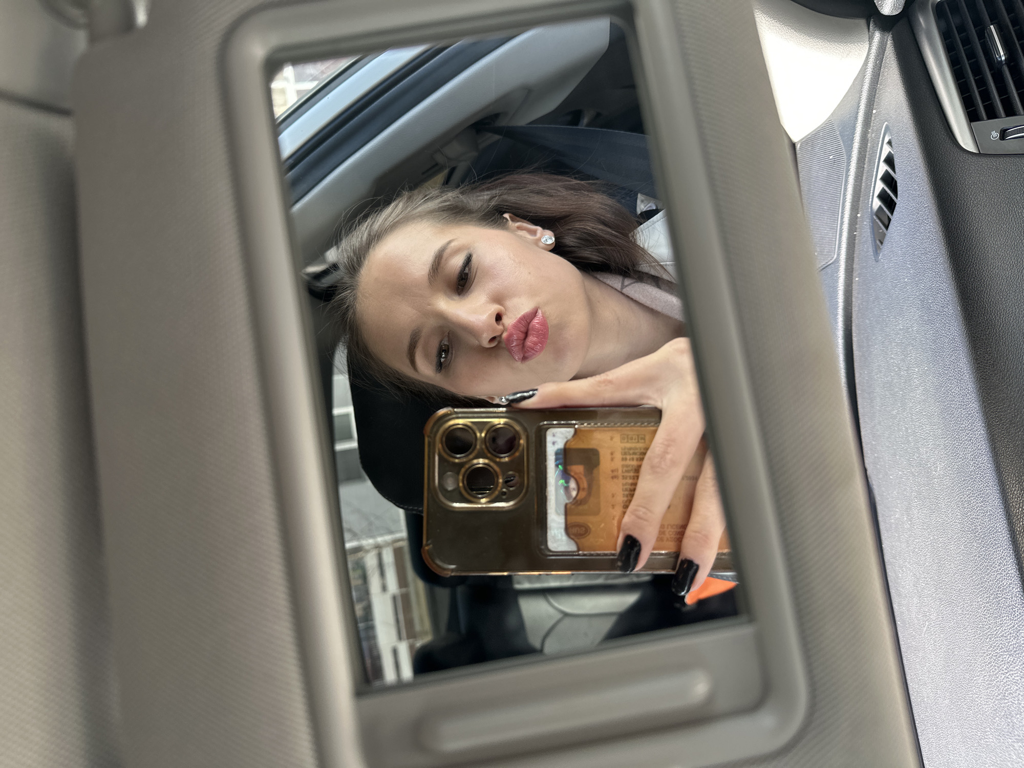 A young woman is taking a selfie in the mirror of the sun visor of a car. The background shows buildings, suggesting an urban setting. The photo was taken in Yekaterinburg, Russia. She is holding a gold iPhone with a card in the case. Her nails are painted black. She is pouting her lips.