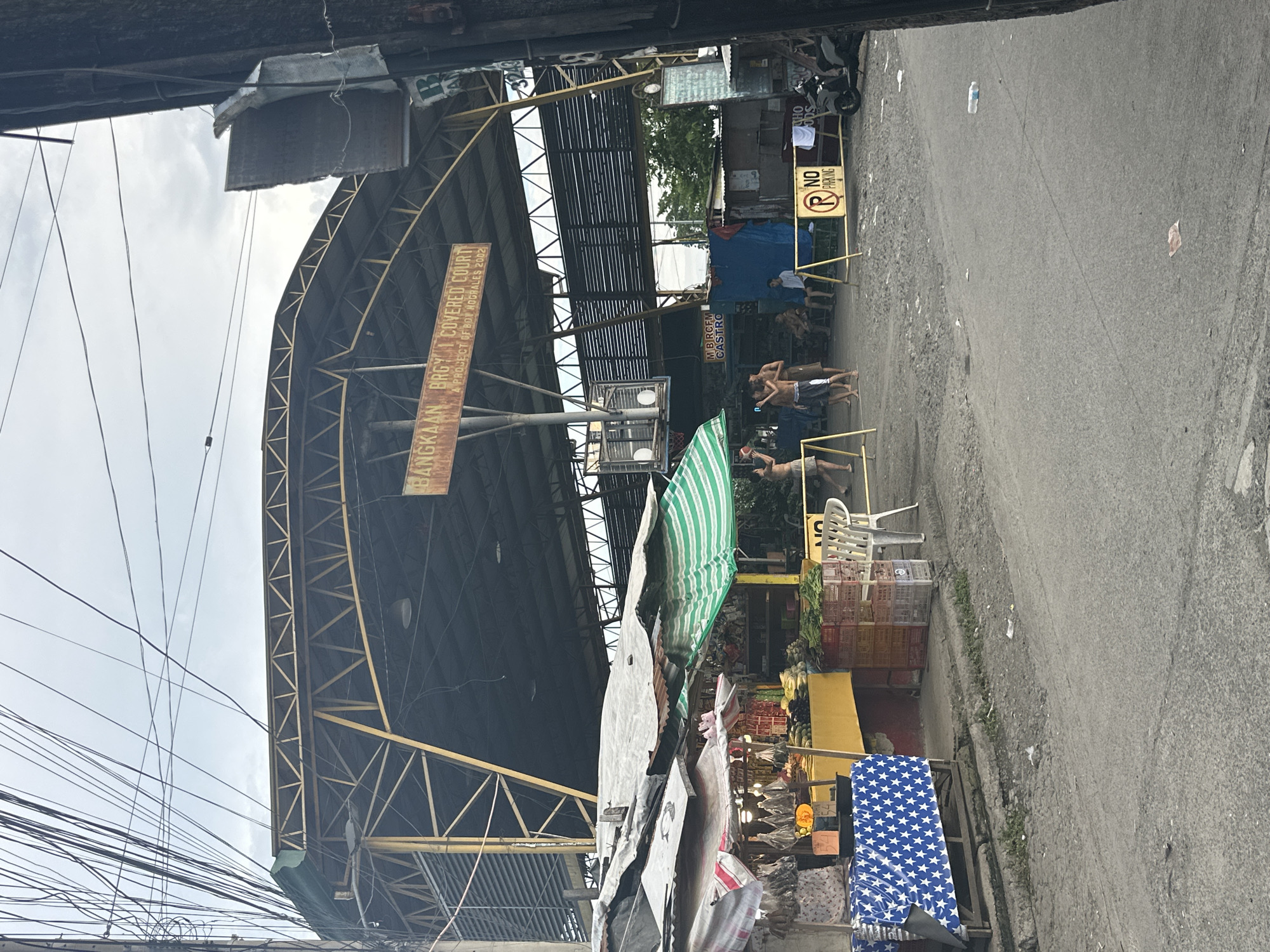 The image captures a scene at the Bangkaan Brgy. 1 Covered Court in Davao City, Philippines, which also functions as a public market. A large, covered structure with a yellow steel frame and a dark roof dominates the background, identified by a weathered sign that reads "BANGKAAN BRGY. 1 COVERED COURT A PROJECT OF BOY NOGRALES 2002".
In the foreground, makeshift market stalls are set up. One stall on the left displays various items, including what appear to be dried goods and produce, covered by a patterned blue and white cloth. Another stall to the right features fresh vegetables, with crates stacked behind it. Several people are present in the scene. A group of young men are visible near the court, some appearing to be playing basketball, while others are seated or standing. A "NO PARKING" sign is prominently displayed, with a motorcycle parked nearby. The ground is unpaved and appears somewhat littered. The overall environment suggests a bustling, community-oriented space that combines recreational and commercial activities, typical of a public market setting in the Philippines. The sky is overcast, indicating cloudy weather.