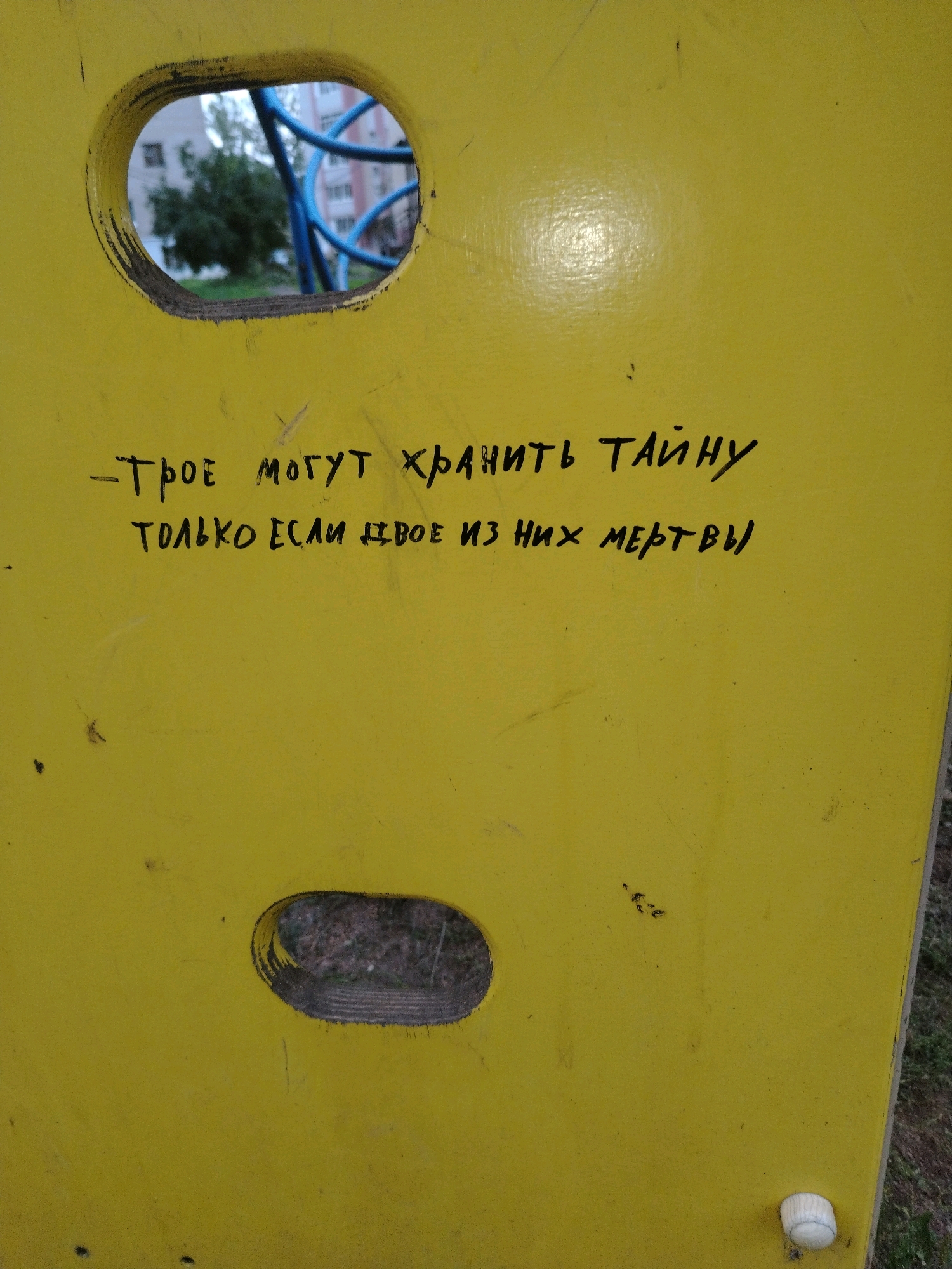 The image is a close-up shot of a yellow, weathered playground structure, likely made of painted metal or plastic. The surface is scratched and scuffed, indicating age and use. There are two oval-shaped cutouts in the structure, through which a glimpse of the surrounding environment can be seen. In the upper cutout, blurry blue pipes and green foliage are visible, suggesting the presence of playground equipment and trees. The lower cutout shows a patch of dry grass and dirt.
Handwritten in black marker on the yellow surface are two lines of Russian text. The first line reads: "Трое могут хранить тайну" which translates to "Three can keep a secret". The second line reads: "ТОЛЬКО ЕСЛИ ДВОЕ ИЗ НИХ МЕРТВЫ" which translates to "Only if two of them are dead".
The image does not contain any identifiable people or specific landmarks that would allow for precise localization within Yaroslavl, Russia. The context suggests an outdoor playground setting in a residential area.