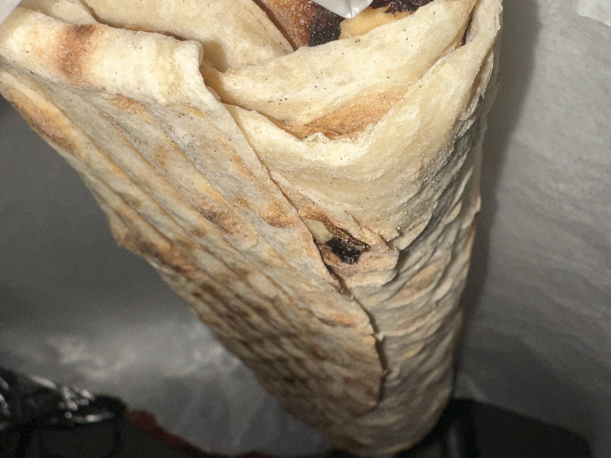 The image displays a close-up of a tightly rolled food item, resembling a wrap, burrito, or a filled flatbread, presented vertically. The wrap is made from a light-colored, thin flatbread or pastry dough, which appears to be lightly toasted or grilled, showcasing subtle brown and darker, slightly charred areas on its surface. The dough exhibits a somewhat textured, layered, and slightly wrinkled appearance due to being rolled. Small, dark specks, possibly seasoning like pepper or herbs, are visible on the surface of the wrap. Towards the interior of the roll and within some folds, glimpses of a dark-colored filling or ingredient can be seen. The food item is partially held or resting against a soft, light-grey or white material, likely a paper bag or a takeaway wrapper, which forms an indistinct background. The lighting suggests an indoor setting, and no specific activities, people, or discernible text are visible. The image captures the texture and details of the food item in close proximity, suggesting it has been recently prepared or purchased in Chaikovsky, Russia.