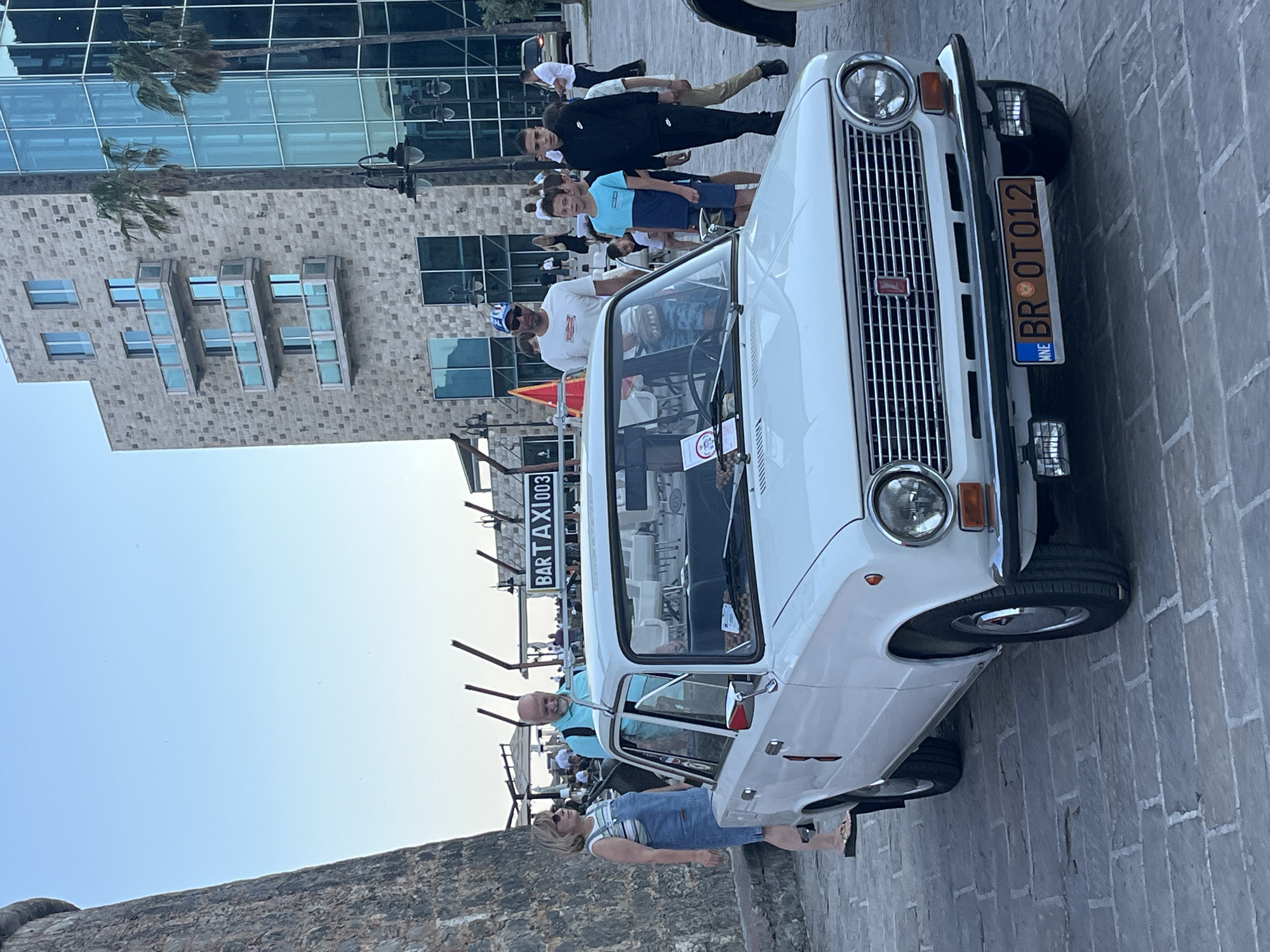 The image shows a white vintage car, identified by the license plate "BR OT012" and potentially a Lada model, parked on a brick-paved area, likely a pedestrian zone. The presence of the "BARTAXI003" sign suggests the location may be near a bar or restaurant. A group of people is standing around the car and in the background, including a woman wearing overalls and sunglasses, who appears to be near the car's door. Behind the people and the car, there is a modern glass and stone building and palm trees, suggesting the location is likely in a tourist area. Given the license plate code "BR", this photo was taken in the city of Budva, Montenegro.
