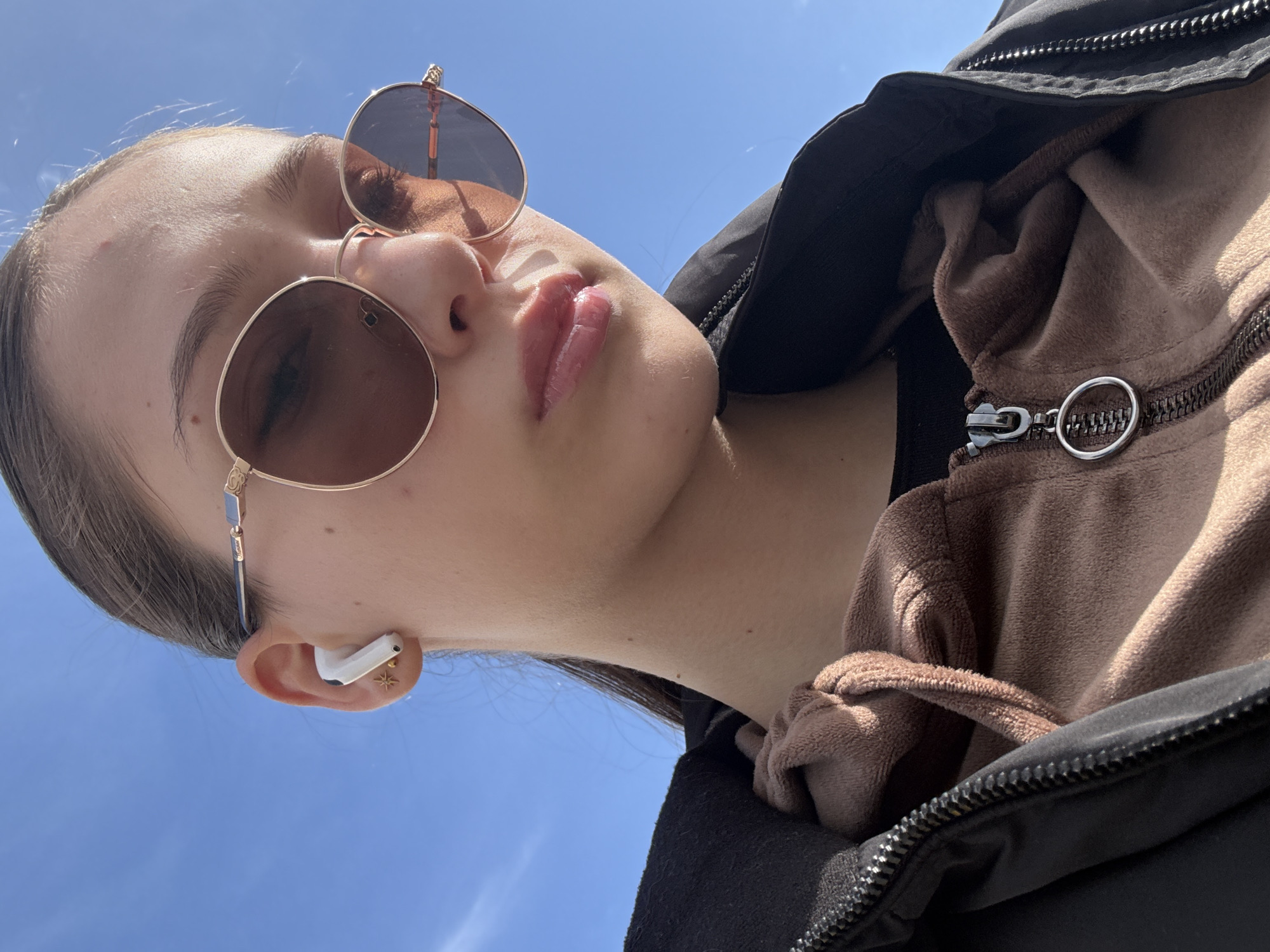 A young woman is photographed from a low angle, looking down towards the camera. She is wearing round sunglasses with gold frames. In her left ear, she has a white wireless earbud and a gold star-shaped earring. She is wearing a brown fuzzy hoodie under a black jacket. The background is a clear blue sky. The location cannot be determined from the image without additional information.