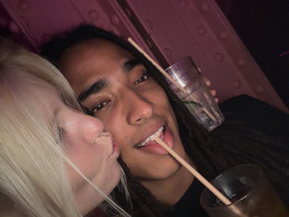 A close-up image reveals a fair-skinned woman with blonde hair kissing a dark-skinned man on the cheek. The man has dreadlocks and a straw playfully protruding from his mouth. He holds two glasses filled with beverages, each containing a straw. The background features a pinkish hue with circular patterns.