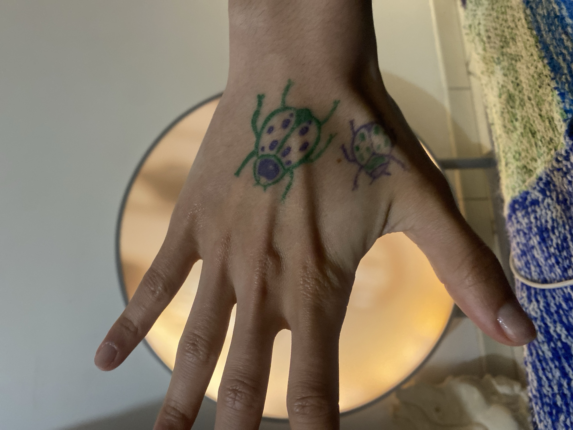 A hand with two drawn ladybugs is displayed prominently. The larger ladybug is drawn in green with dark purple spots and head, while the smaller one is drawn in purple with dark purple spots. The hand is fair-skinned with visible veins and wrinkles. The background features a light-colored surface on the left and a round, illuminated object behind the hand. To the right, a blue and green knitted item is partially visible. There is no information in the image to determine that the image was taken in Aubervilliers, France.