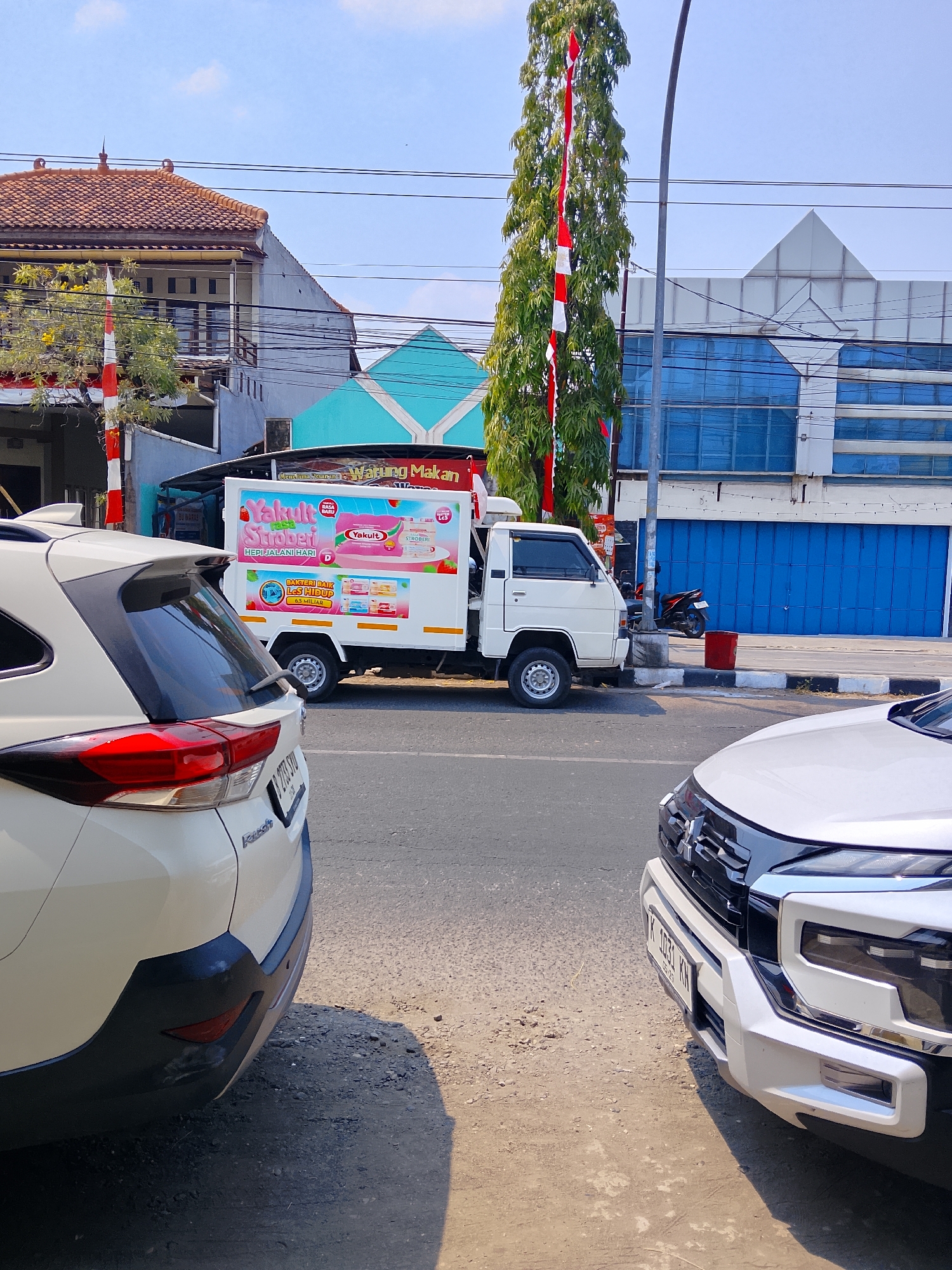 The image depicts a street scene in the city of Ngelo, Indonesia, on a sunny day. In the foreground, the rear of a white SUV is visible on the left, and the front of another white vehicle is on the right. Between the two vehicles, a white delivery truck with "Yakult" branding is parked. The truck has advertisements for Yakult Strawberry and "Bakteri Baik Les Hidup 6.5 Miliar" (Good Bacteria Live Life 6.5 Billion) on its side. Behind the truck, a building with a turquoise roof and white geometric patterns is partially visible. Further in the background, there are other buildings with tiled roofs and a larger modern building with blue glass windows. A tall tree decorated with Indonesian flags (red and white stripes) stands near the street. A motorcycle is parked in front of a building with blue rolling shutters. The street is paved, and the ground beside the road is dusty and gravelly. The sky is clear with some light clouds.