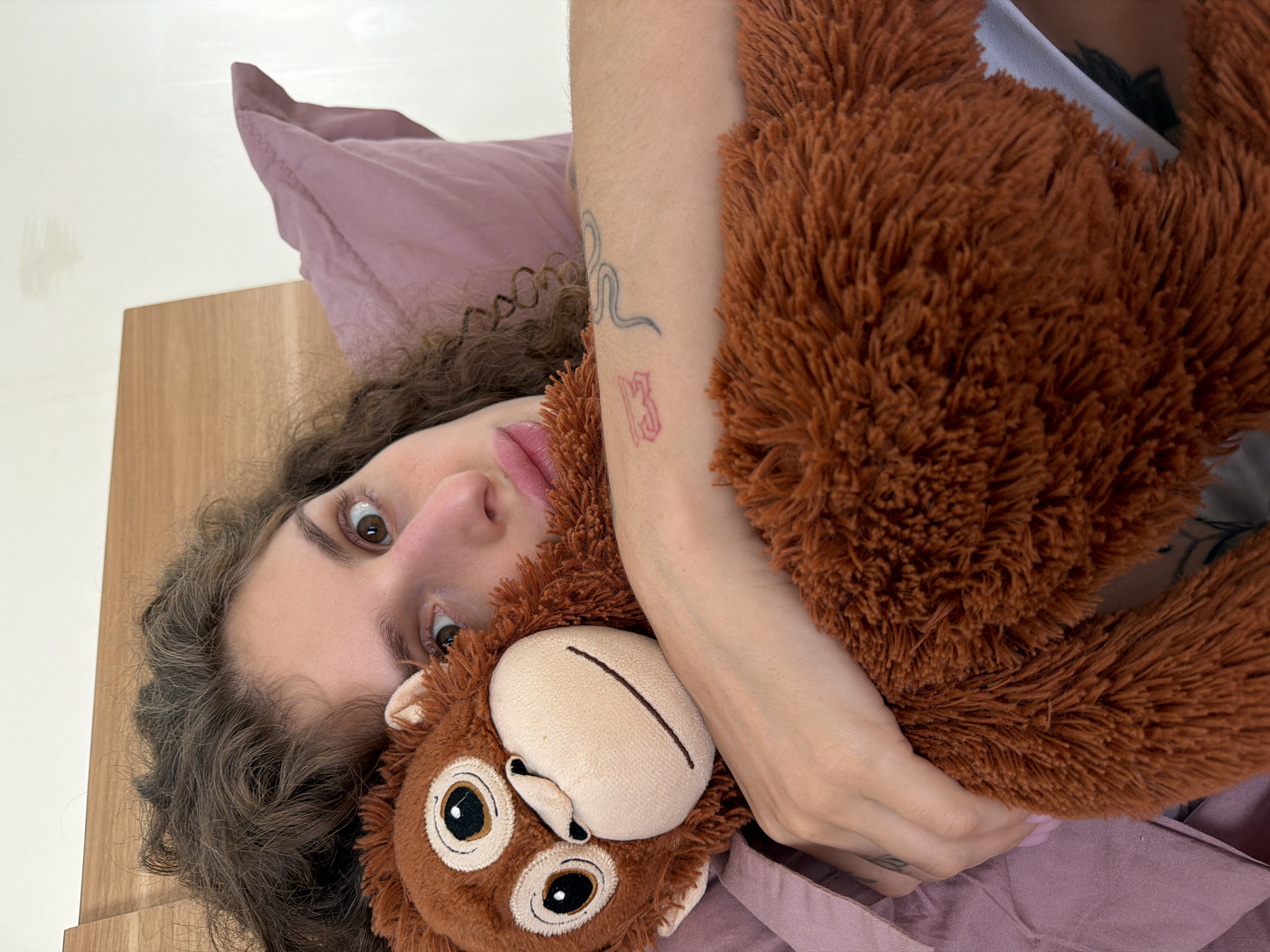 A person is holding a stuffed monkey while lying in bed. The person has curly brown hair and is looking at the camera. The stuffed monkey is brown with a tan face. The person is lying on a pink pillow and is covered with a pink sheet. The background is a white wall and a brown wooden cabinet. The location is unknown, and cannot be determined from the image.