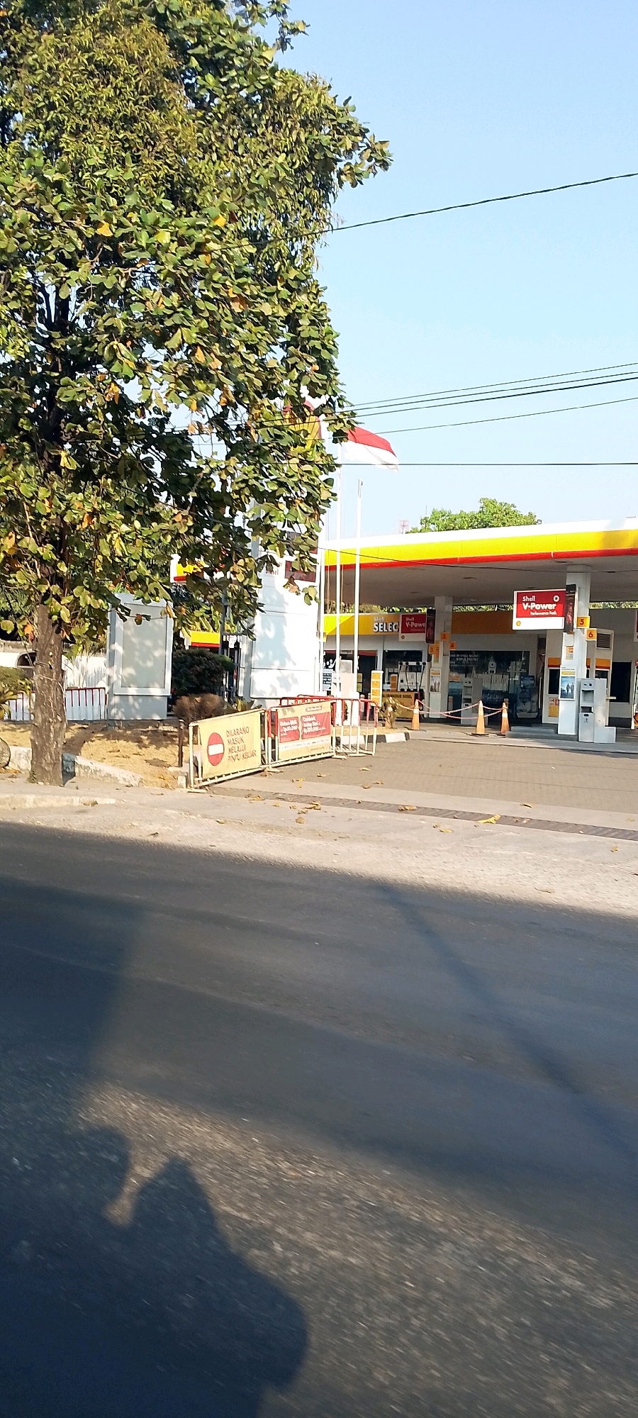 The image captures a section of a Shell V-Power gas station in Bekasi, Indonesia, under a clear blue sky. A large tree with green and yellowing leaves partially obscures the view of the station. The gas station features its signature yellow and red canopy, with several pumps visible. A prominent sign reading "SELECT V-Power" is displayed. A red and white Indonesian flag flutters from a pole on the station's roof. In the foreground, a road surface is visible, with shadows cast across it, suggesting it is daytime. Yellow barriers with warning signs are positioned near the entrance of the gas station, one of which displays text in Indonesian that appears to be a prohibition. The overall environment suggests a typical urban setting in Indonesia during daylight hours.
