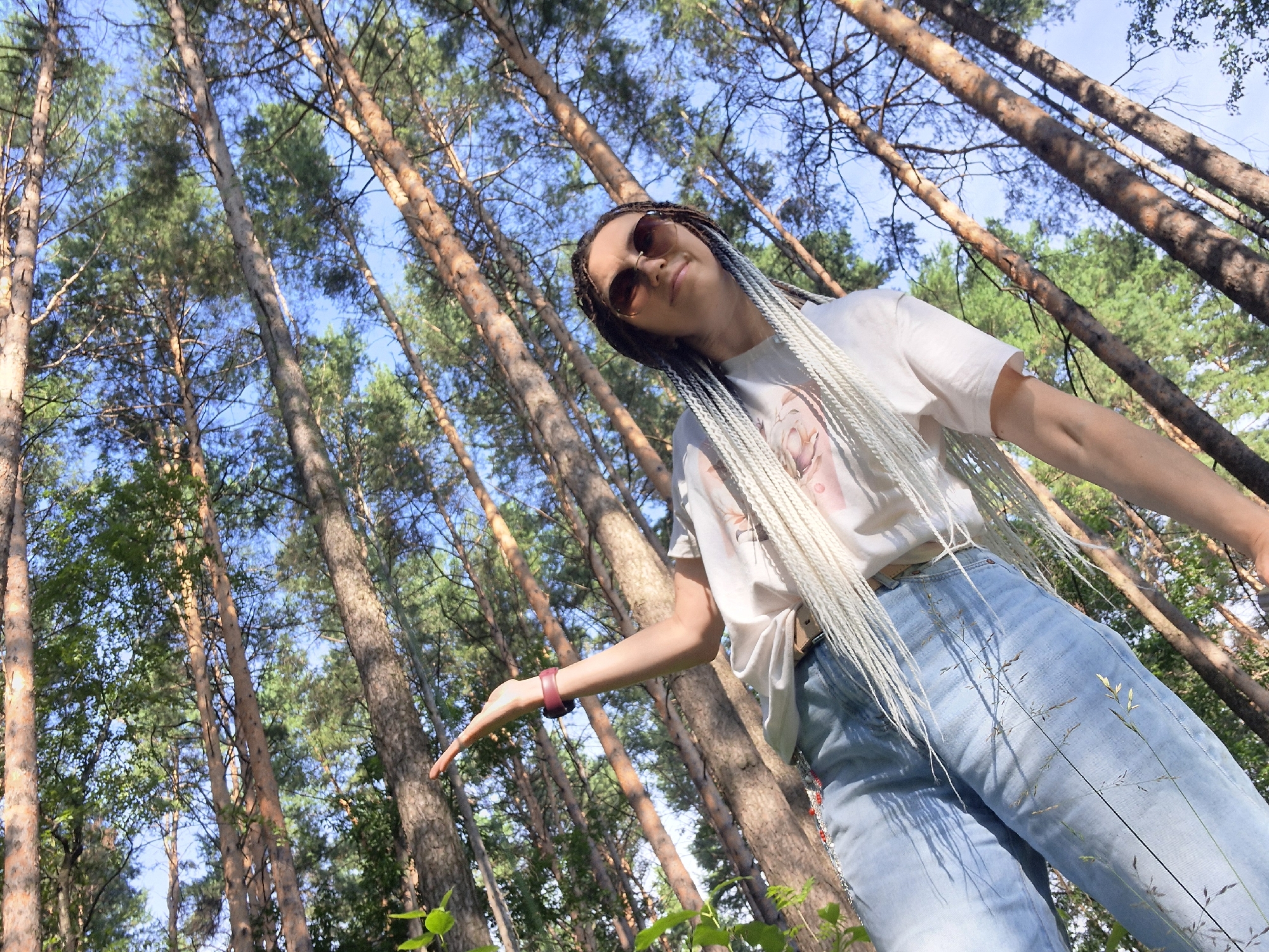 A low-angle, full-shot photograph captures a young woman with long blonde braids posing in a forest. She wears light blue jeans, a white t-shirt with a printed design, and sunglasses. Her left arm is raised with her palm open, and she wears a dark red fitness tracker on her wrist. The background is dominated by tall pine trees with a clear blue sky visible through their branches. The image appears to be taken in a natural outdoor setting, possibly a park or wooded area within the city of Chaikovsky, Russia, given the presence of pine trees common in the region. The lighting suggests it is daytime, with sunlight filtering through the trees, casting dappled shadows.