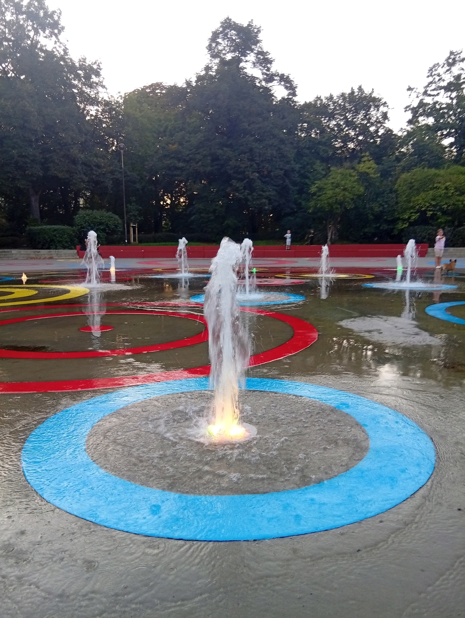 The image depicts a splash pad in Wrocław, Poland, likely during daylight hours. Several fountains are active, spraying water into the air. The ground is wet, with colorful concentric circles painted on it, featuring blue, red, and yellow. In the background, there are trees and a red seating area. A few children are visible, one standing near the seating area and another in the water. A dog is also present near the seating area, interacting with a person. The overall environment suggests a public park or recreational area designed for children.