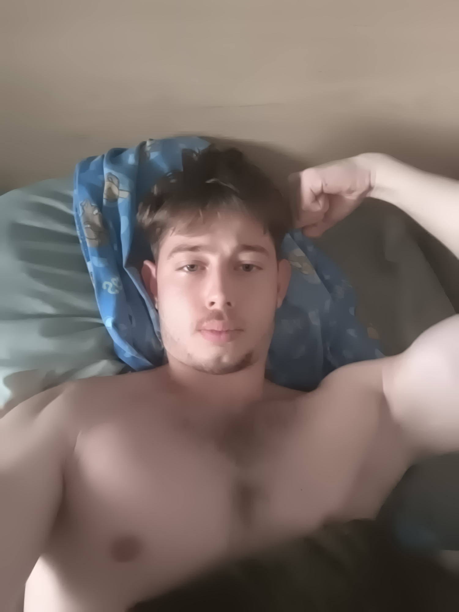 A young adult male, likely in his 20s, is shown in a close-up, chest-up selfie while lying down indoors. He has short, light brown hair, a light mustache, and a small goatee. He is shirtless, displaying a muscular physique, with his left arm bent and fist clenched near his head, and his right arm also bent, showcasing his bicep.
He rests on a bed, with his head supported by a blue pillow adorned with cartoon characters, possibly resembling Scooby-Doo. Another plain blue or teal pillow or sheet is visible beneath the patterned one, and darker bedding is present on the right side of the frame. The background consists of a plain, light-colored wall.
The man looks directly into the camera with a neutral to slightly serious expression. The lighting is soft and ambient, characteristic of an indoor setting with indirect light. No specific time of day or weather conditions can be discerned, and there are no visible location cues within the image itself to indicate Sofia, Bulgaria. No text is visible in the media file.