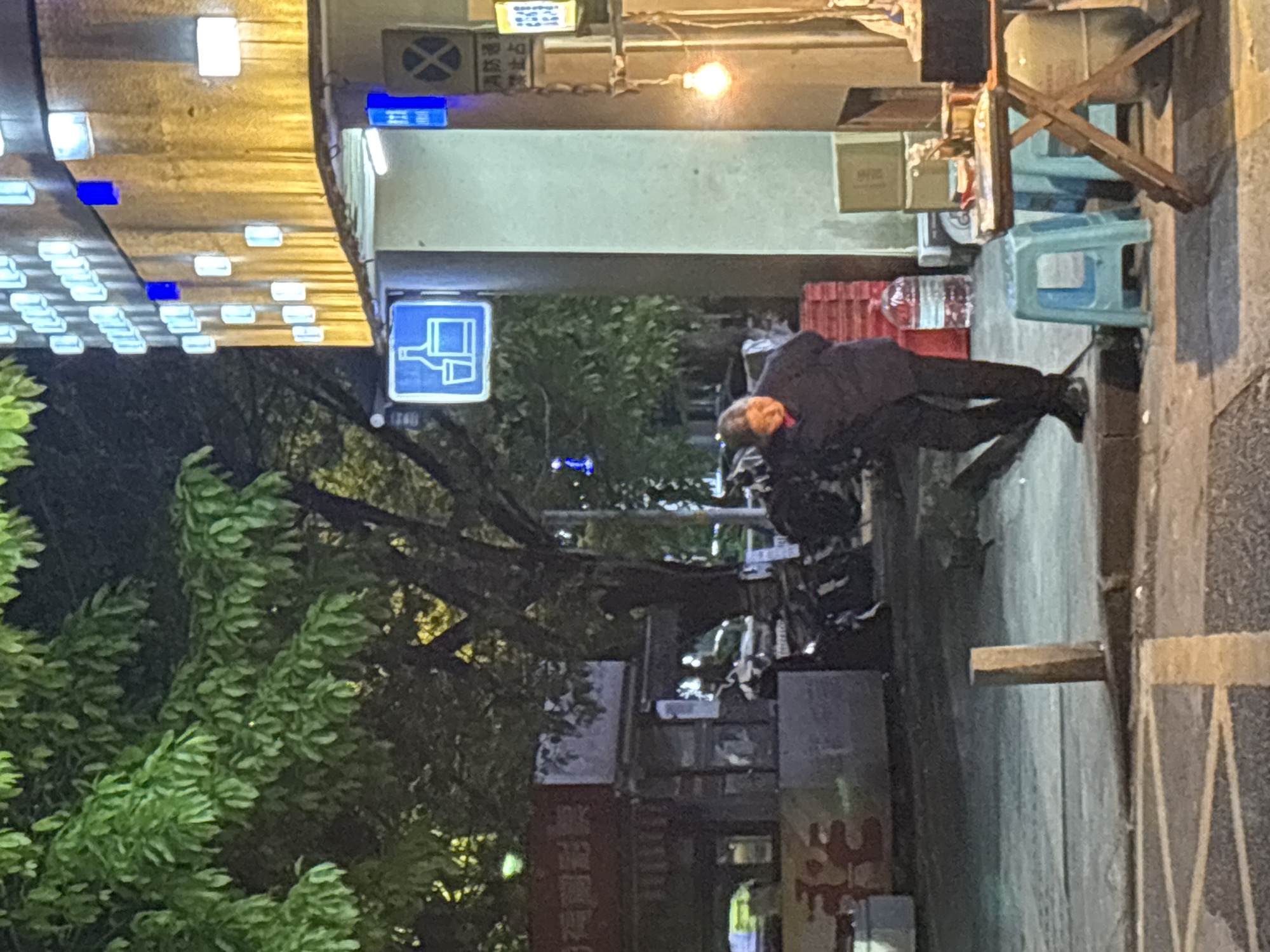 The image depicts an urban scene, possibly in Nan'an District, China. The environment is a street at night, with elements like streetlights, trees, and signs. A man in dark clothing is visible, possibly resting or leaning against something. There are stools and crates along the street, potentially indicating a vendor or street food setup. Buildings line the street and there's evidence of commercial activity. The overall setting is a bustling urban area at night.