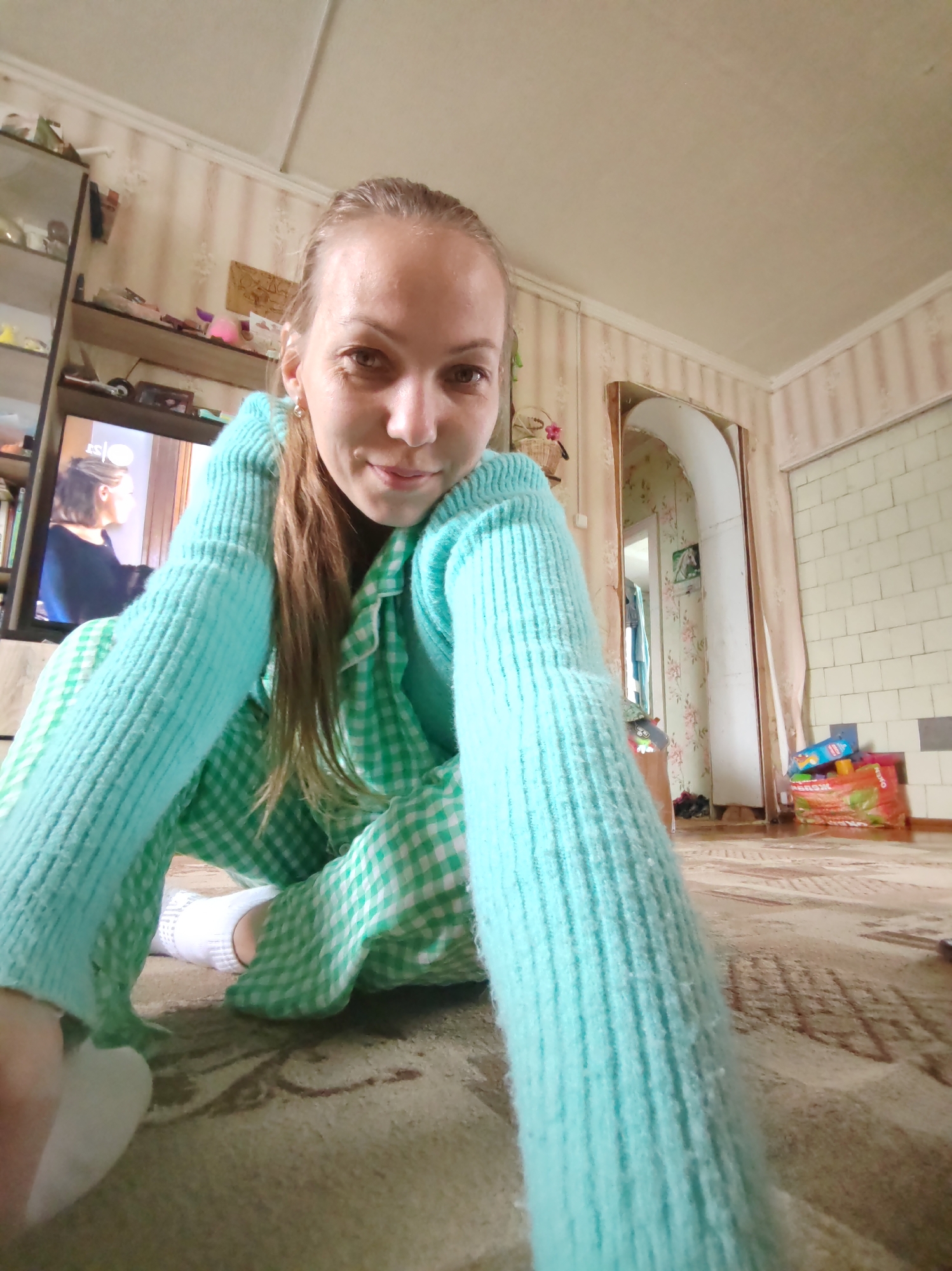 The image shows a young woman in pajamas, sitting on a carpeted floor. She is wearing a green and white checkered pajama set and a light blue knitted sweater. Her hair is pulled back, and she is looking at the camera with a slight smile.
In the background, there is a television screen displaying an image of a woman. To the left of the television, a shelf unit is visible, filled with books and various objects. The room has patterned wallpaper and a doorway leading to another area of the house. In the far right, a pile of colorful boxes and bags can be seen.
The overall impression is of a casual, indoor setting, likely a home. The lighting suggests it could be daytime. The woman appears relaxed and comfortable, taking a selfie.