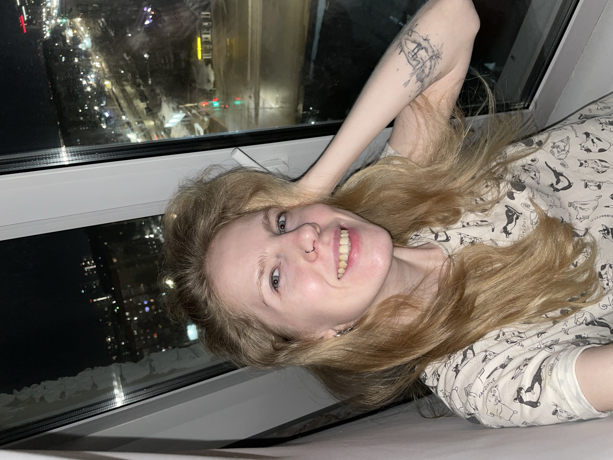 A young, light-skinned woman with blond hair and blue eyes is lying on a white surface near a window. She is smiling, revealing her teeth and a nose ring. She has a tattoo on her left arm. The woman is wearing a light-colored shirt with a cat pattern. Outside the window, a cityscape is visible at night, with numerous lights indicating a populated area. Based on visual cues and the overall composition of the image, the location is likely Izhevsk, Russia. The environment suggests an urban setting at night.