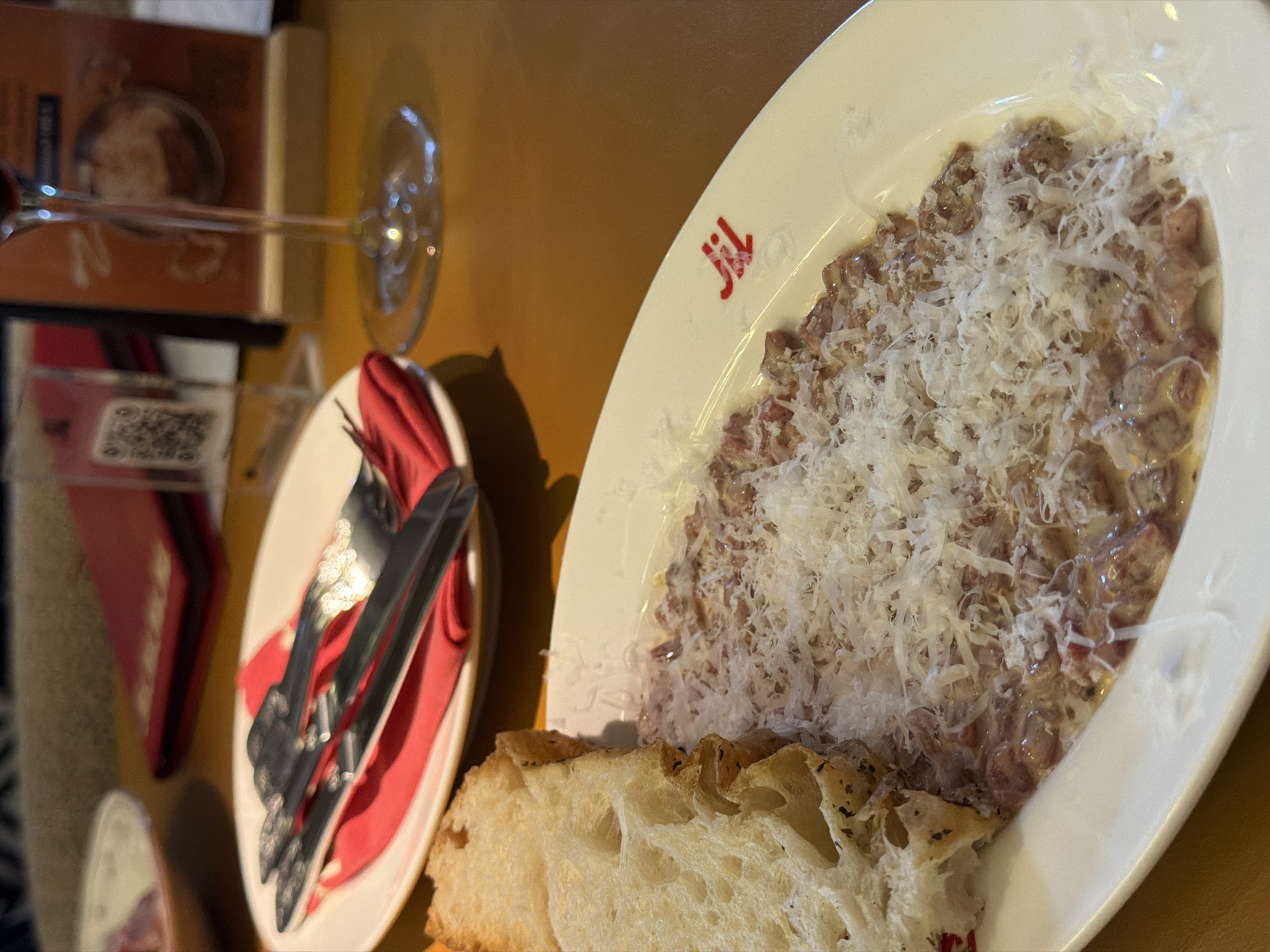 This image presents a close-up view of a dining table, likely in a restaurant or cafe, illuminated by warm, artificial light. The central subject is a white, oval-shaped plate filled with a creamy, reddish-brown dish, generously topped with finely grated white cheese, resembling Parmesan. A piece of rustic, light-colored bread, possibly focaccia, with visible air pockets and some seasoning, rests against the plate. A distinct red logo, appearing as "Jil," is visible on the rim of the main plate.
To the left, a smaller white plate holds a neatly folded red napkin, upon which a fork and knife are placed. Further back and to the left, a clear wine glass stands, reflecting ambient light. In the background, out of focus elements include a red menu holder or card displaying a QR code, and a blurred brown booklet or menu with some text and an image. The table surface is a warm, light brown tone. The overall scene suggests a meal in progress, with no people visible. The photograph is taken from a slightly elevated, angled perspective.