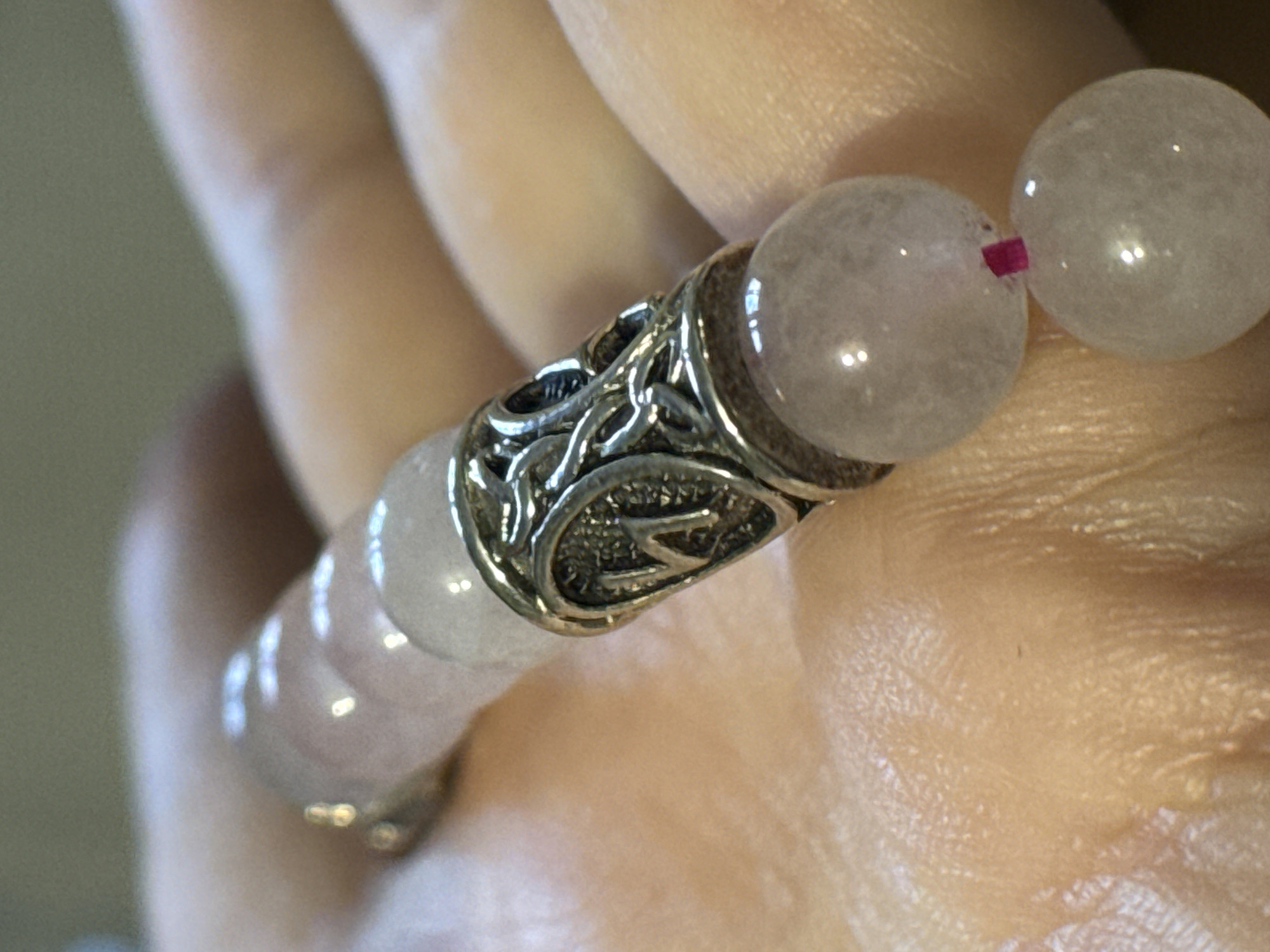 A close-up image shows a pink beaded bracelet with a silver ornate cylindrical spacer held in a human hand. The beads appear to be rose quartz. A pink cord threads between two beads. The silver spacer has a textured surface with a letter 'Z' design on one side. The skin of the hand is visible with pores and wrinkles, suggesting an older person. No details indicate the location of Novo-Nikol'skoe, Russia.