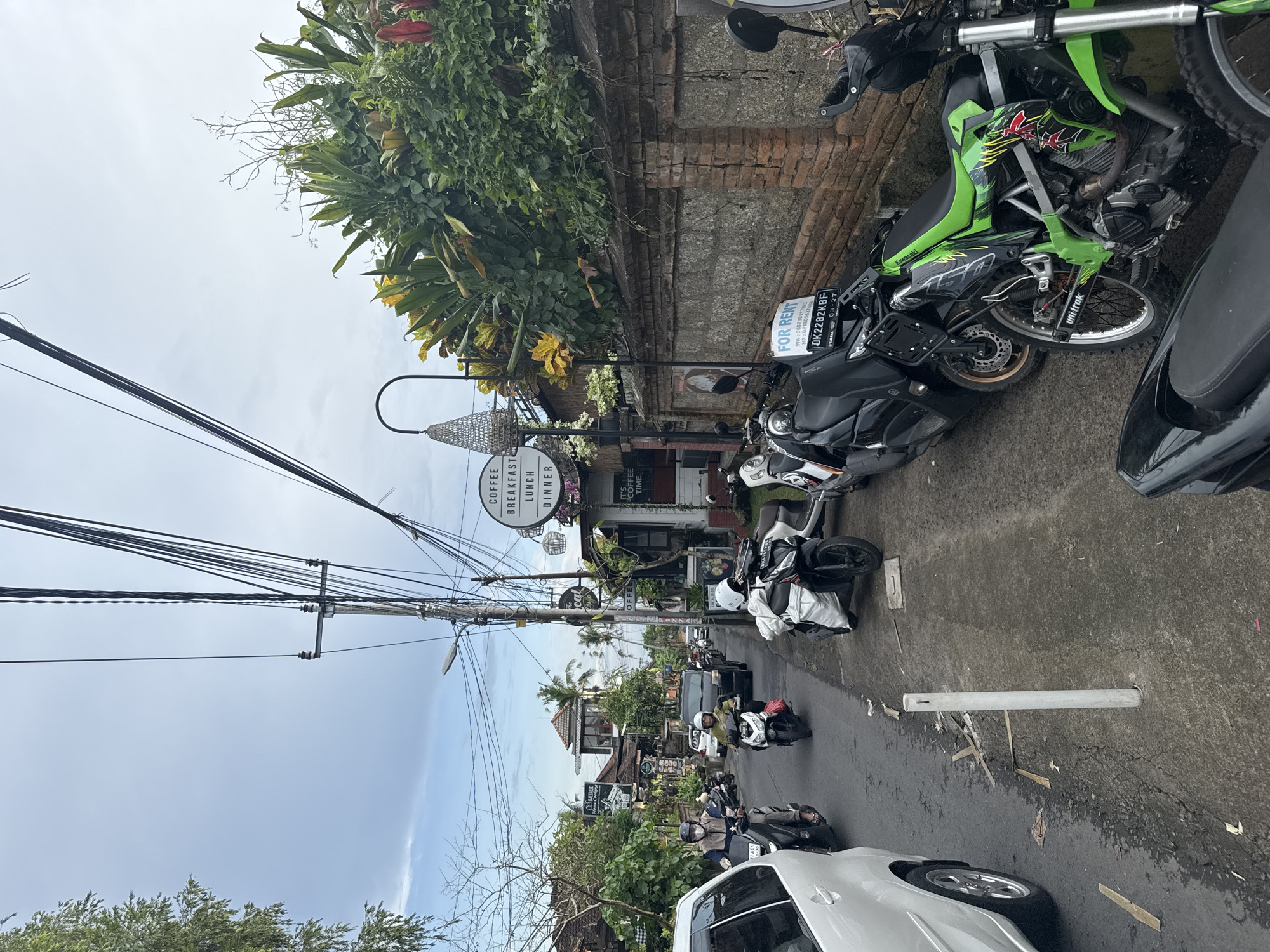 This image captures a vibrant street scene, likely in a tropical setting such as Ubud, Indonesia, during the day. The perspective is from the side of the road, looking down a street lined with buildings and dense tropical foliage.
The main subjects include numerous motorcycles and scooters, both parked and in motion, dominating the streetscape. A prominent green Kawasaki dirt bike is parked on the right, displaying "unitrak" on its suspension. Several black scooters are also parked, one bearing a "FOR RENT" sign. A white car is partially visible in the foreground on the left. Individuals are seen riding scooters further down the road, some wearing helmets.
Along the street, various buildings house businesses. A round sign clearly advertises "COFFEE BREAKFAST LUNCH DINNER," with a smaller sign below stating "IT'S COFFEE TIME." Another "COFFEE" sign is also visible. The architecture features brick walls and some traditional-style roofs, blending with modern elements. Lush green and yellowing foliage, along with some red flowers, adorn the buildings and roadside, contributing to a verdant atmosphere.
Overhead, a complex network of utility poles and numerous power lines crisscrosses the sky, a common feature in many urban areas of Southeast Asia. The sky is bright but largely overcast, suggesting it's either mid-morning or early afternoon. The activity depicts a typical day with traffic movement and open businesses.
Visible text includes: "COFFEE BREAKFAST LUNCH DINNER," "IT'S COFFEE TIME," "FOR RENT," "WA: 085738117993," "HP: 0818056374," "DK2282KBF," "03-27," "Kawasaki," and "unitrak."