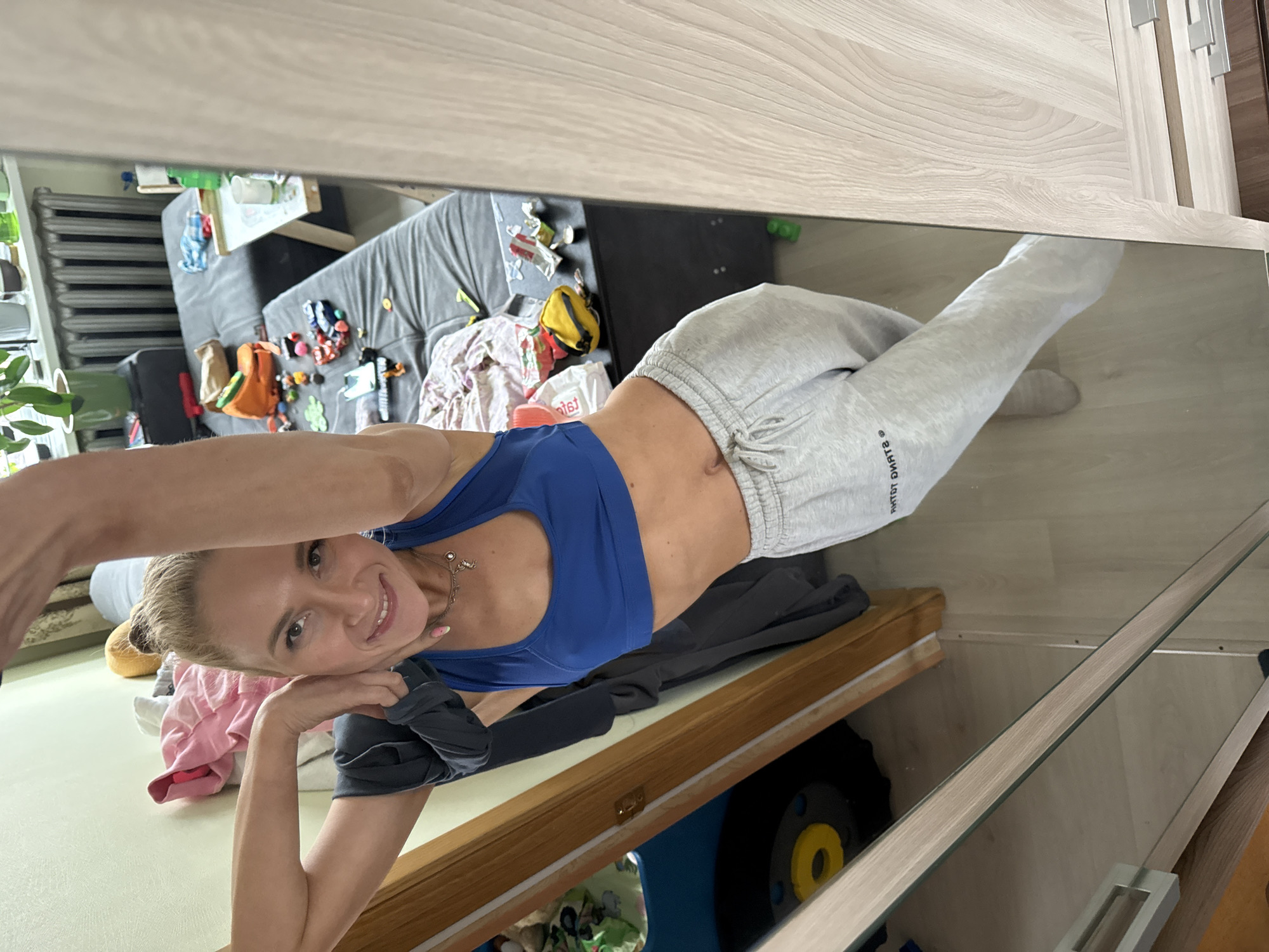 The image displays a rotated mirror selfie taken by a young woman in an indoor setting in Chaikovsky, Russia. The woman is visible in her reflection, positioned horizontally across the frame, smiling warmly at the camera. She wears a blue sports bra and light grey sweatpants, with the text "SPORT SKATE" visible on her left pant leg. A delicate necklace is around her neck.
The reflection behind her reveals a casual room, likely a living room or bedroom, with a large, grey upholstered couch. The couch is notably cluttered with various items, including colorful toys, clothing, a yellow bag, and what appears to be cups and food wrappers. A grey radiator is visible further back in the reflected room.
In the actual foreground, above the mirror, there is a wooden surface, possibly a shelf or part of a wardrobe. Below this surface, to the woman's left, some folded clothes (including a pink shirt) are visible. Further down, partially obscured by the frame, is a black object with a yellow circular detail, possibly exercise equipment or a toy. The overall lighting suggests it is daytime. The image appears to be shot from a slightly elevated perspective, looking down into the mirror.