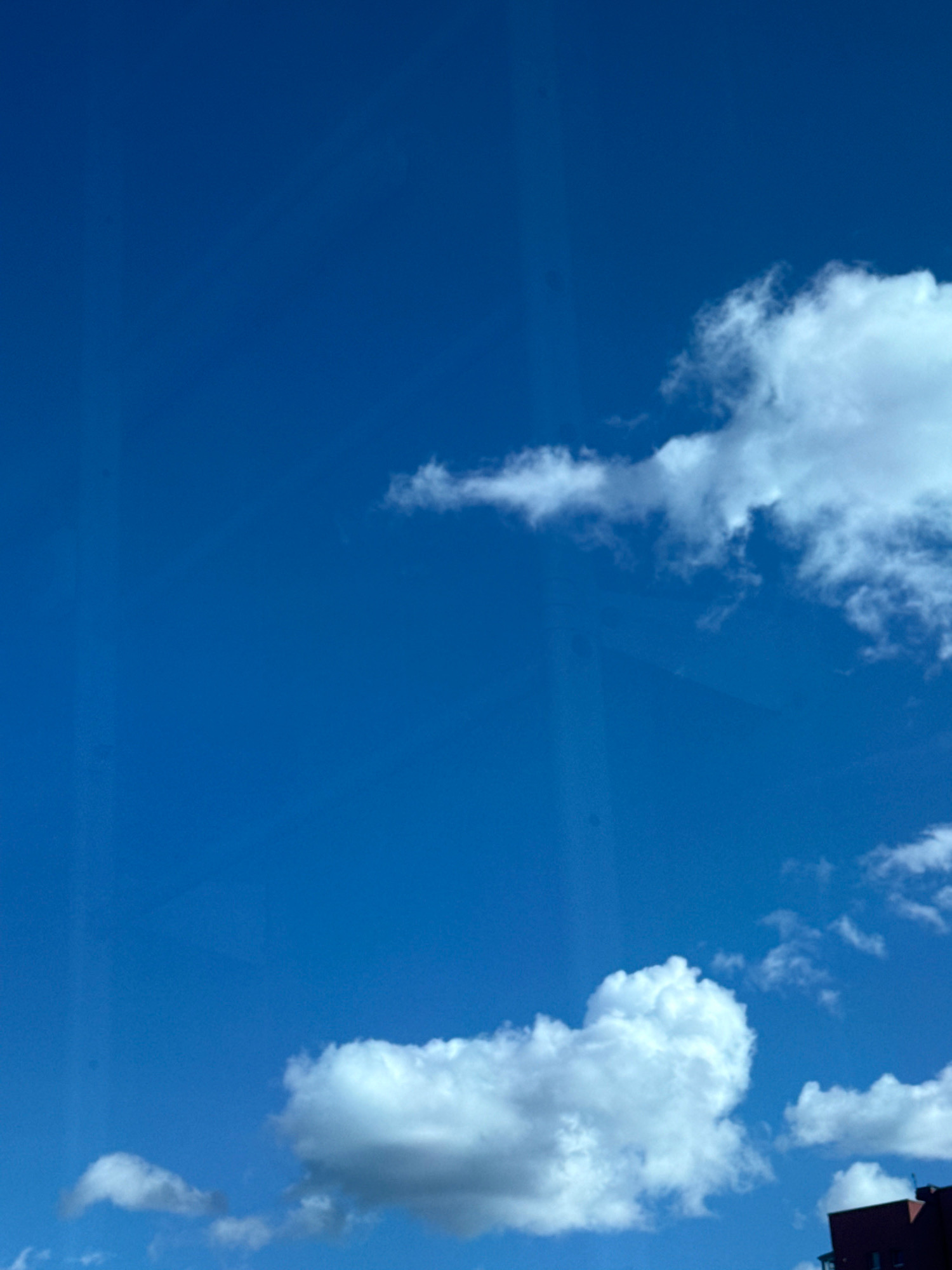 The image shows a clear blue sky with scattered white cumulus clouds. In the lower right corner, a dark red building with dark windows is partially visible, suggesting an urban setting. The building appears to be modern and tall. The sky occupies the majority of the frame, with the clouds creating a dynamic contrast against the deep blue. There are no people or any discernible actions taking place. The overall impression is of a sunny day in an urban environment, likely in Tallinn, Estonia, given the architectural style of the building.