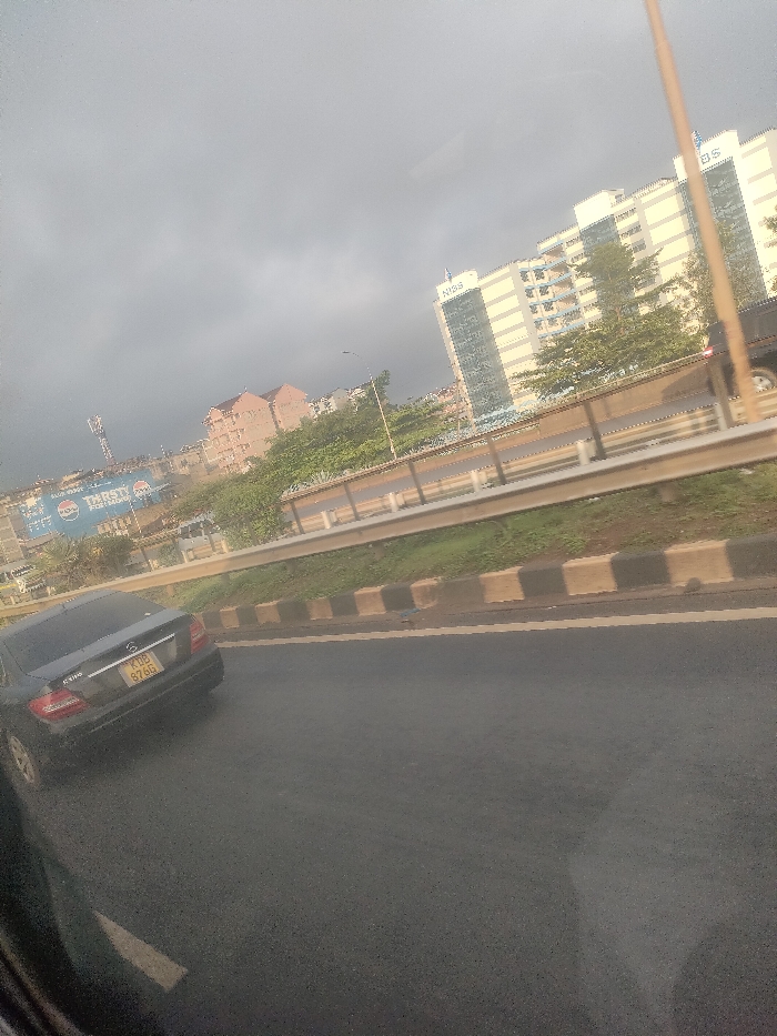 The image depicts an urban street scene under an overcast sky, located in Theta ward, Kenya. In the foreground, the lower left quadrant shows a partial view of a vehicle's dark interior frame. The mid-ground features a dark asphalt road with a partial white lane marking on the left. A black Mercedes-Benz C180 sedan, bearing Kenyan license plate KUB 876G, is visible on the road ahead, angled slightly to the right. To the right of the road is a raised concrete median, painted with alternating black and light brown blocks, topped by a strip of grass and a metallic guardrail.
In the background, beyond the median and guardrail, an elevated road or overpass is visible with a vehicle partially seen on its right lane. Lush green trees and vegetation line the area behind the elevated structure. Several buildings are present, most notably a tall, multi-story building with light-colored facades and blue-tinted windows, featuring the "NISS" logo at its summit. Adjacent to it, another similar building displays "KISS" signage. Further to the left and rear, multiple smaller buildings with reddish-brown tiled roofs and lighter facades are visible. A large blue billboard featuring the Pepsi logo and the text "THIRSTY PREMIUM" is prominent on the left, partially obscured by a tall communications tower. No individuals are distinctly visible.