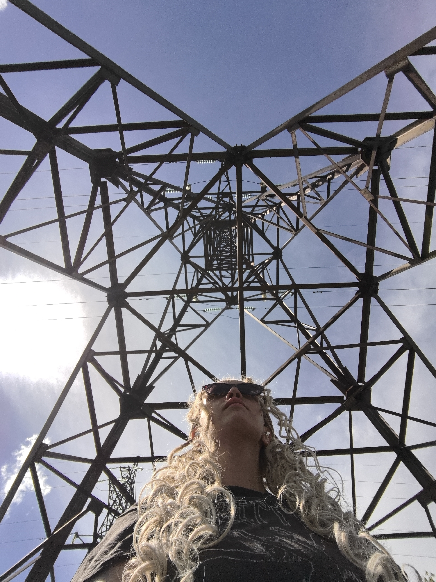 A low-angle shot shows a person with long, curly blonde hair and sunglasses looking up. They are wearing a dark t-shirt with a graphic design. The person is positioned beneath a large, dark metal power line pylon, viewed from below. The pylon's intricate lattice structure extends upwards and outwards, forming a geometric pattern against a bright blue sky. Sunlight streams from the left, creating a lens flare and highlighting some clouds. The perspective emphasizes the scale of the pylon and the clear sky. The scene appears to be outdoors, possibly in a rural or industrial area, given the presence of the power lines. The time of day is daytime, indicated by the bright sun and clear sky.