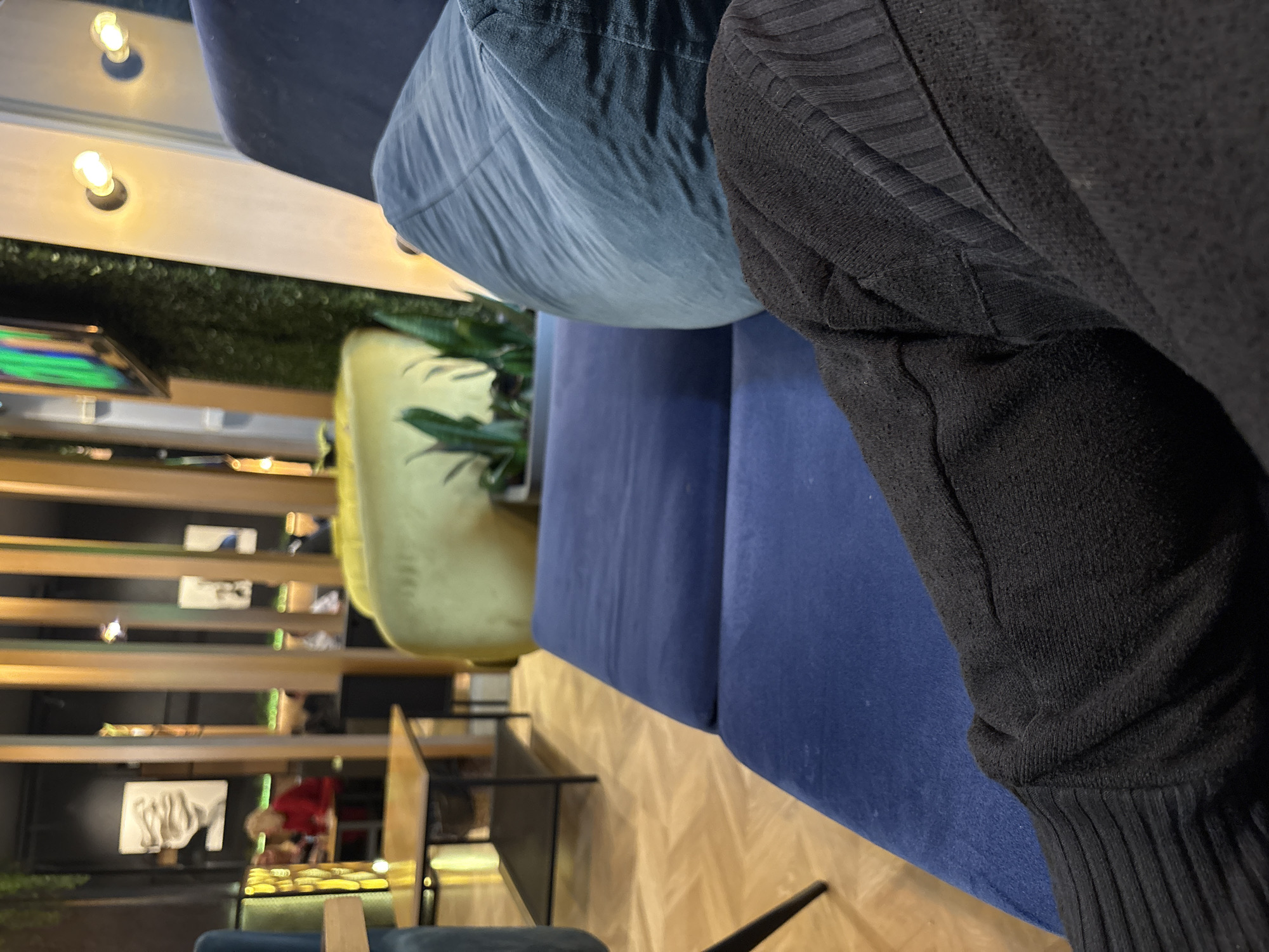 The scene depicts the interior of a modern café or lounge located in Stara Zagora, Bulgaria, viewed from a seated, first-person perspective. In the immediate foreground, the observer's legs are visible, covered in dark, possibly ribbed or textured fabric, resting on a blue upholstered sofa or bench. A lighter blue cushion or pillow is positioned on the sofa to the observer's right.
The midground features a light-colored wooden floor with a herringbone or parquet pattern. A low, rectangular coffee table with a wooden top and black metal legs is positioned to the left. A blue upholstered chair or sofa is partially visible on the far left. Towards the center-right, a large, yellow upholstered armchair or pouf is present. Several green leafy plants are visible in planters behind the yellow seating and against a textured, dark wall covered in artificial foliage.
The background reveals a dark wall with decorative vertical wooden slats serving as a room divider or design feature. Several wall-mounted light fixtures with exposed, warm-toned bulbs illuminate the space. Abstract artwork, including a white, face-like sculpture or painting, adorns the walls. A television screen displaying green and purple colors is mounted high on the left. Blurred figures of individuals are discernible in the background; one person wearing a red top is seated at a table on the far left. The overall lighting is artificial and warm, consistent with an indoor commercial establishment.