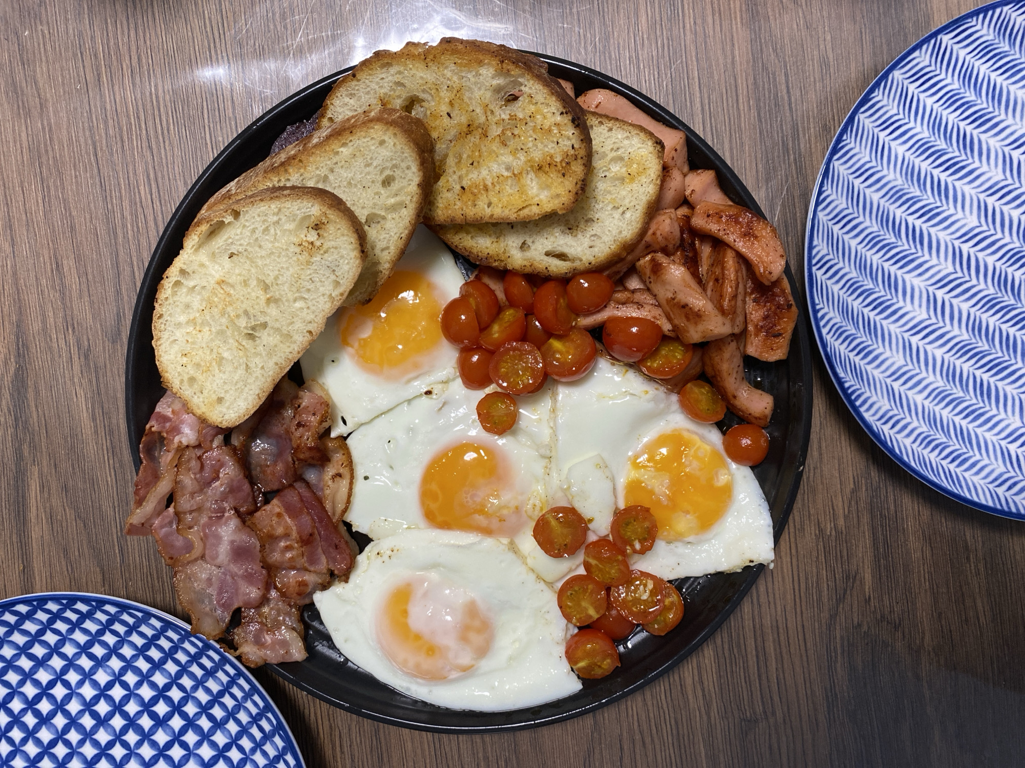 A top-down, medium shot captures a black, circular plate filled with a breakfast meal set on a dark wood-grain table. To the left and bottom right of the plate, portions of blue and white patterned plates are visible. The main plate contains fried eggs with runny yolks, crispy bacon strips, sliced sausages, halved cherry tomatoes, and toasted bread slices. The lighting is even, suggesting an indoor setting. The image is static and shows no people or discernible action. There is no information available about the location in Saint Petersburg, Russia.