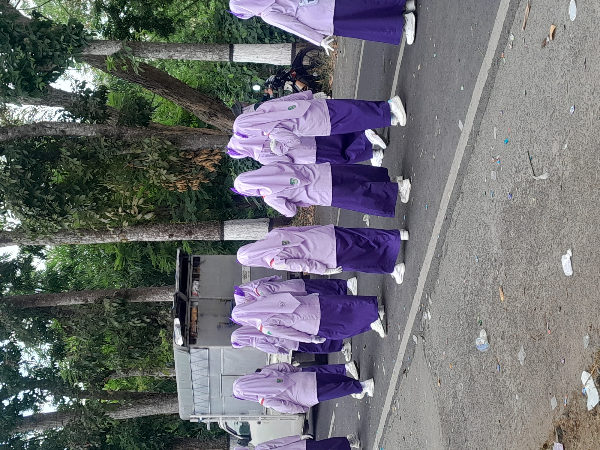 In Sempu, Indonesia, a group of students dressed in purple hijabs and long purple skirts are walking on the side of a road. They are also wearing white gloves and white sneakers. Behind them, a white truck with a tarp covering its cargo is parked. The road is paved and has a white line marking its edge. The surroundings are lined with tall trees with green leaves. Some litter is scattered on the road. A motorcycle is visible in the background.